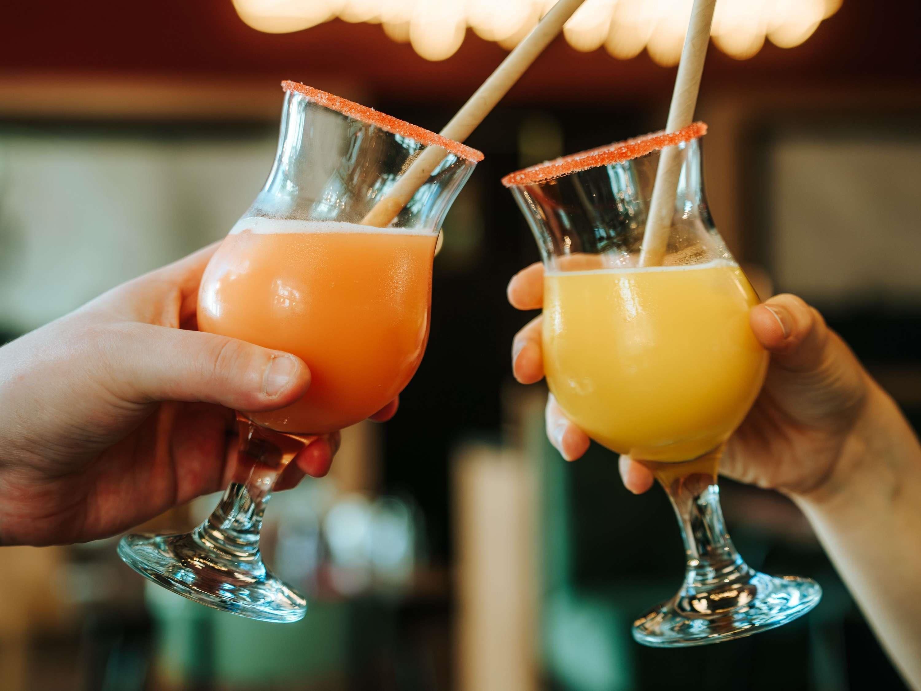 Two people holding glasses with straws, one with an orange drink and one with a yellow drink.