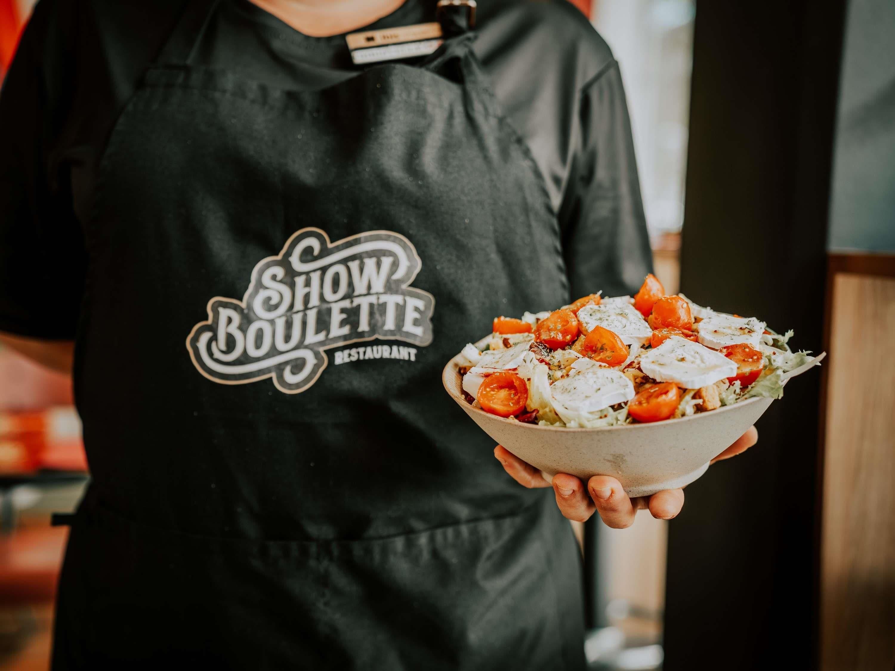 A person holding a bowl of salad with a black apron labeled 'Show Boullette Restaurant.'