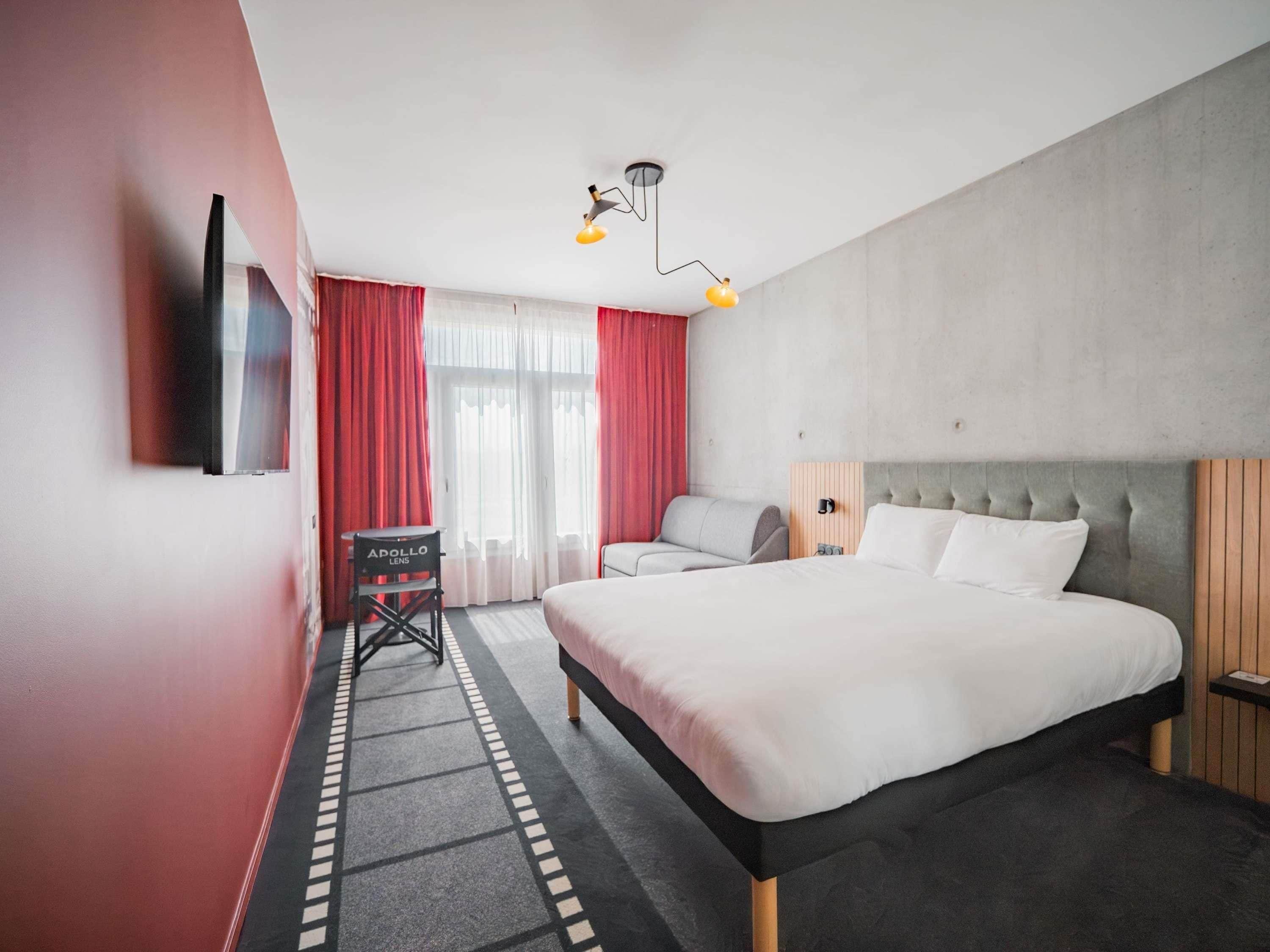 A hotel room with a large bed, a desk, a chair, a TV, and a window with curtains.