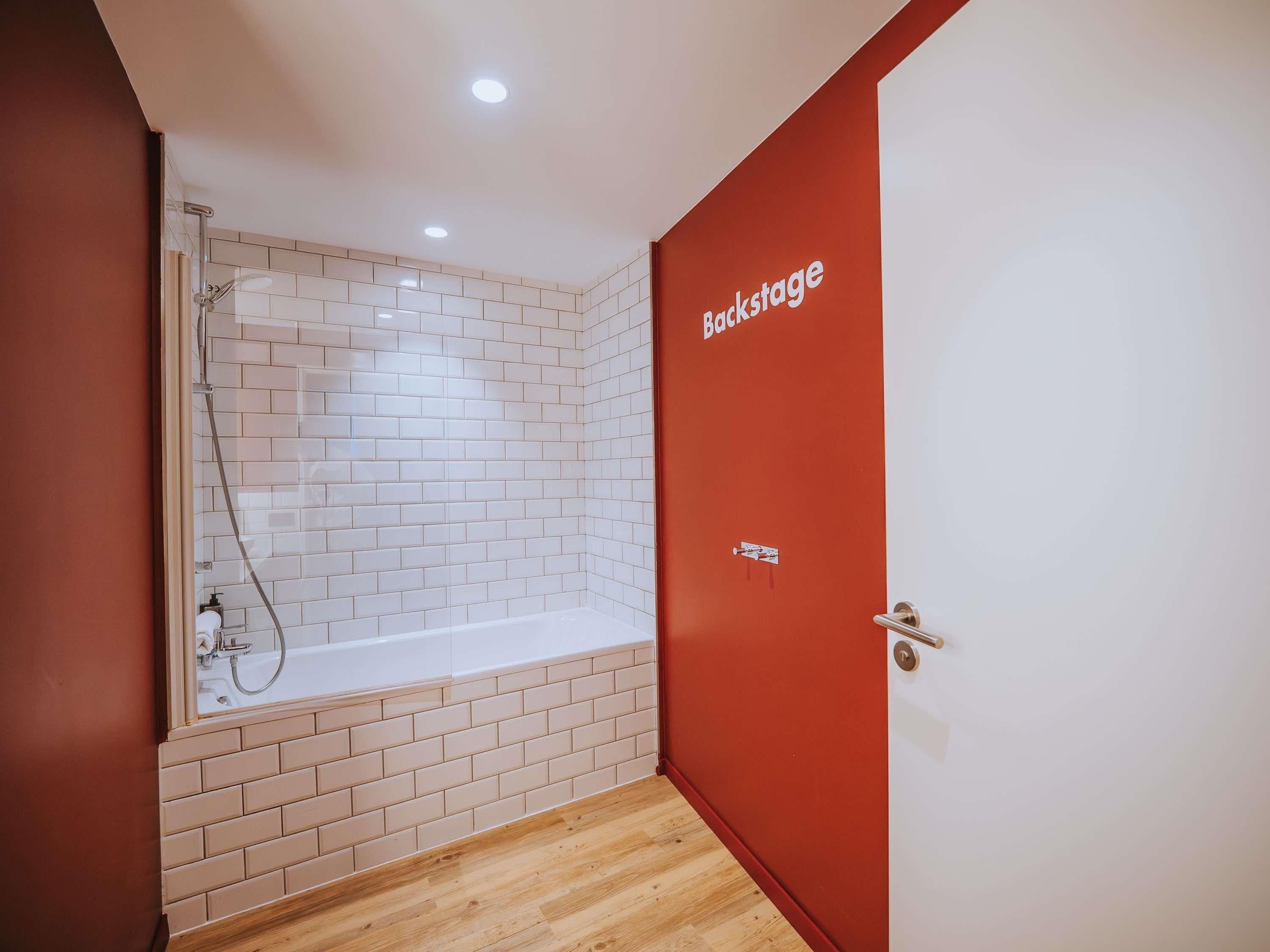 A bathroom with a shower, a bathtub, and a red door with 'Backstage' written on it.
