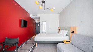 Una habitación de hotel moderna con una pared de acento roja, una cama, un sofá gris, una mesita y un televisor montado en la pared.
