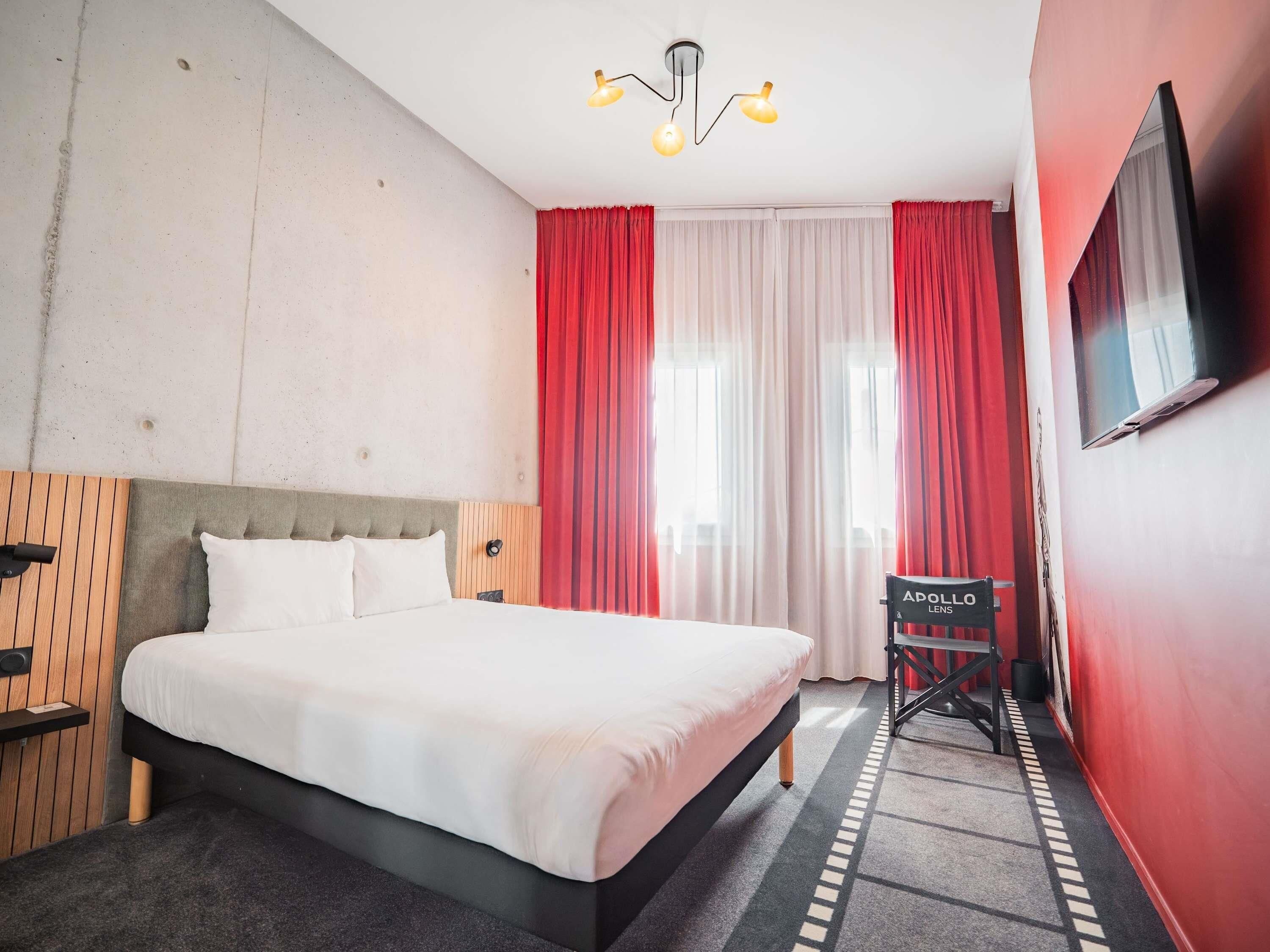 A hotel room with a bed, a TV, a chair, and a window with curtains.