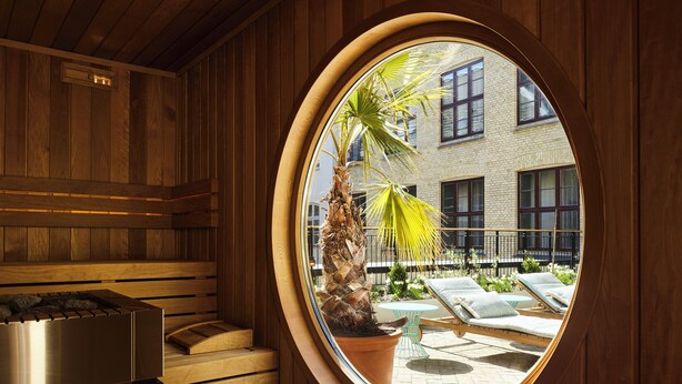 Un sauna con interior de madera, un ventanal redondo y vistas a una zona de estar exterior con una palmera.