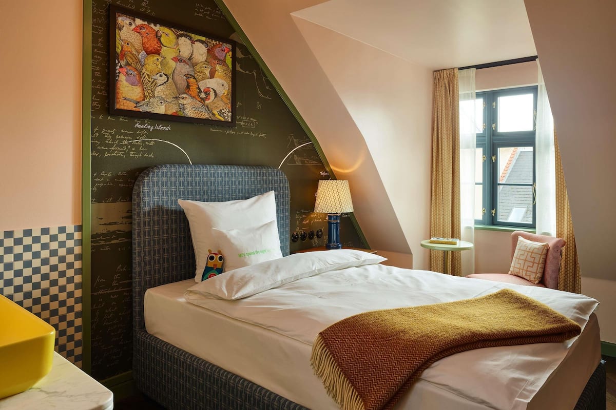 A hotel room with a bed, a pillow with a bird design, a lamp, a window with curtains, and a wall with a chalkboard.