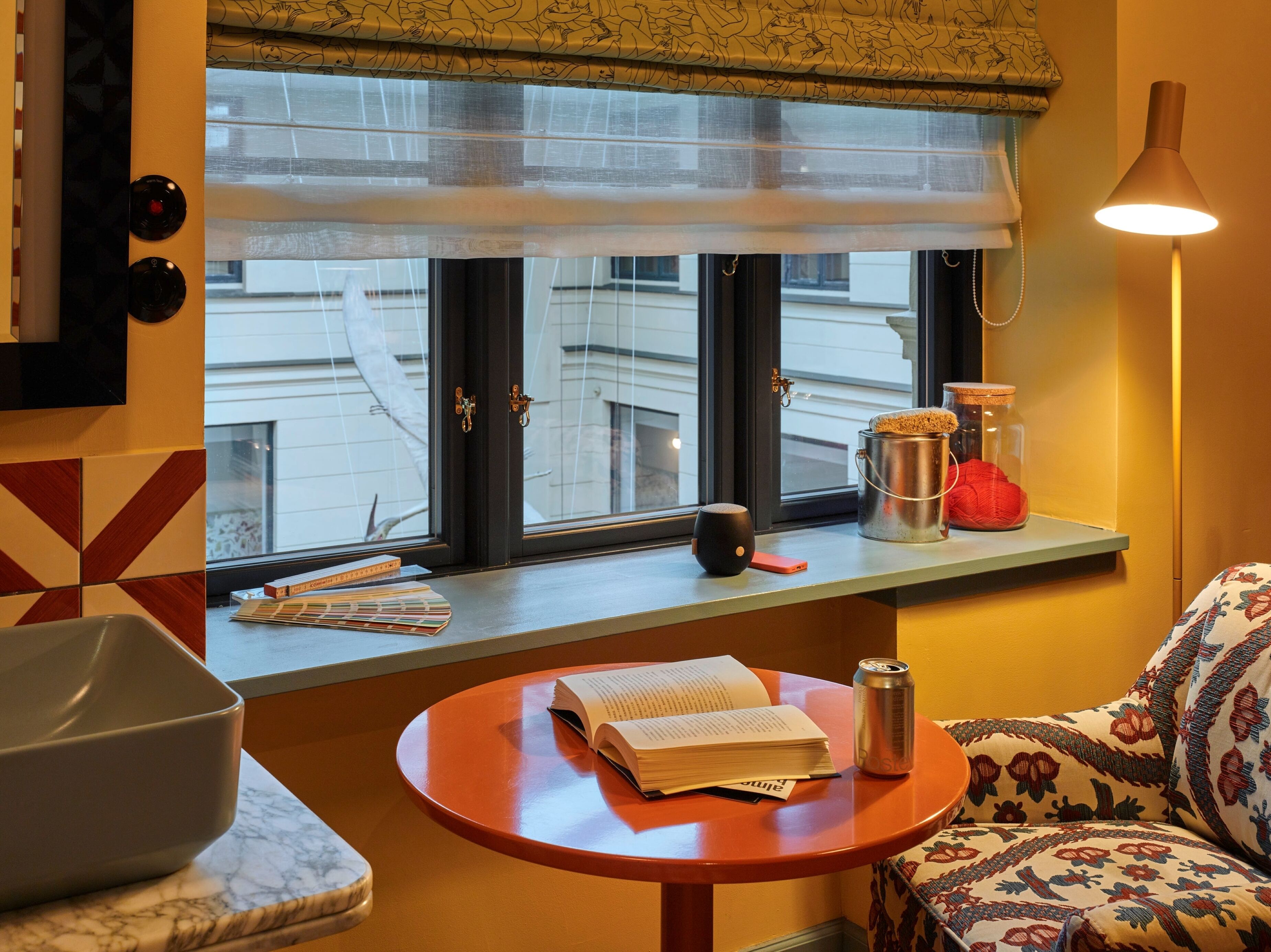 Una habitación con ventana, una mesa con un libro y una lata, una lámpara y un sofá estampado.