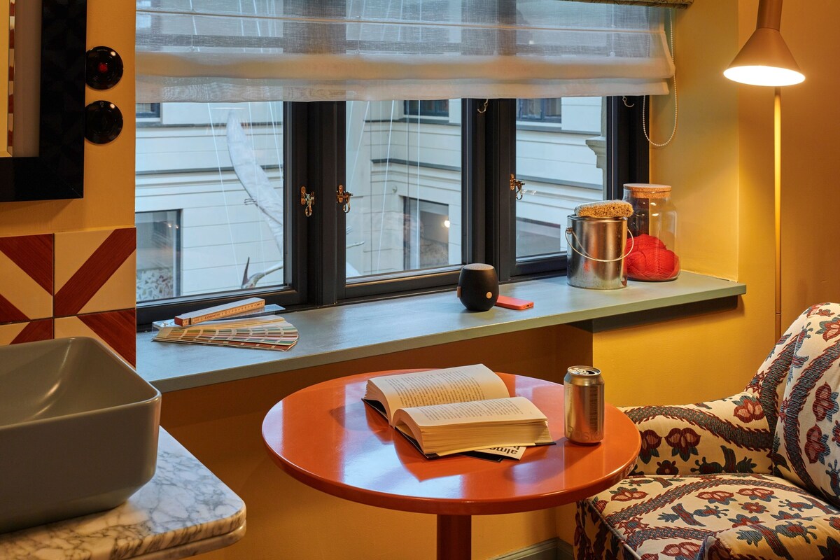 Una habitación con ventana, una mesa con un libro y una lata, una lámpara y un sofá estampado.
