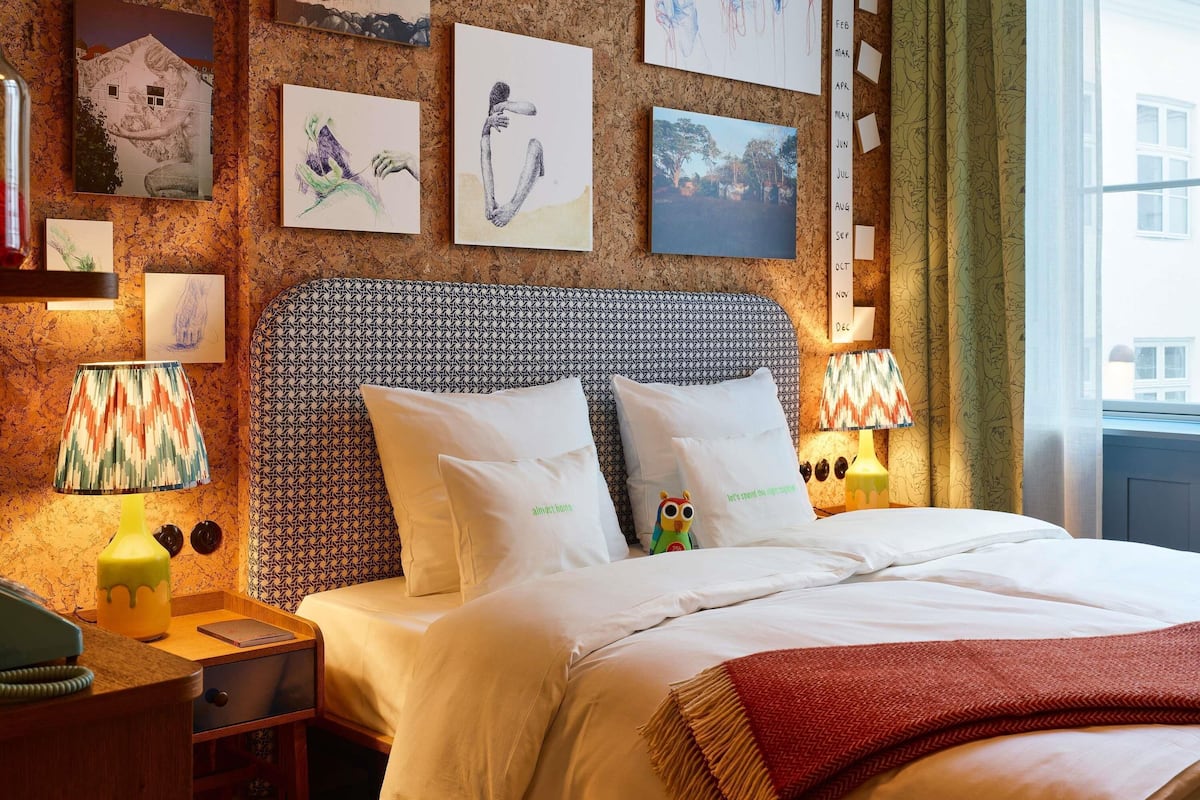 A hotel room with a patterned headboard, a bed with white bedding, a nightstand with a lamp, and artwork on the walls.