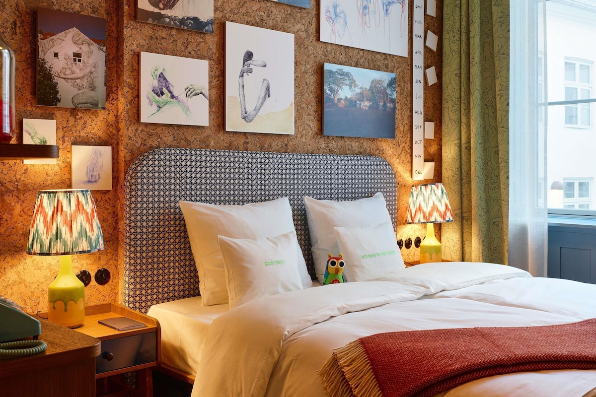 Una habitación de hotel con un cabecero estampado, una cama con ropa de cama blanca, una mesita de noche con lámpara y obras de arte en las paredes.