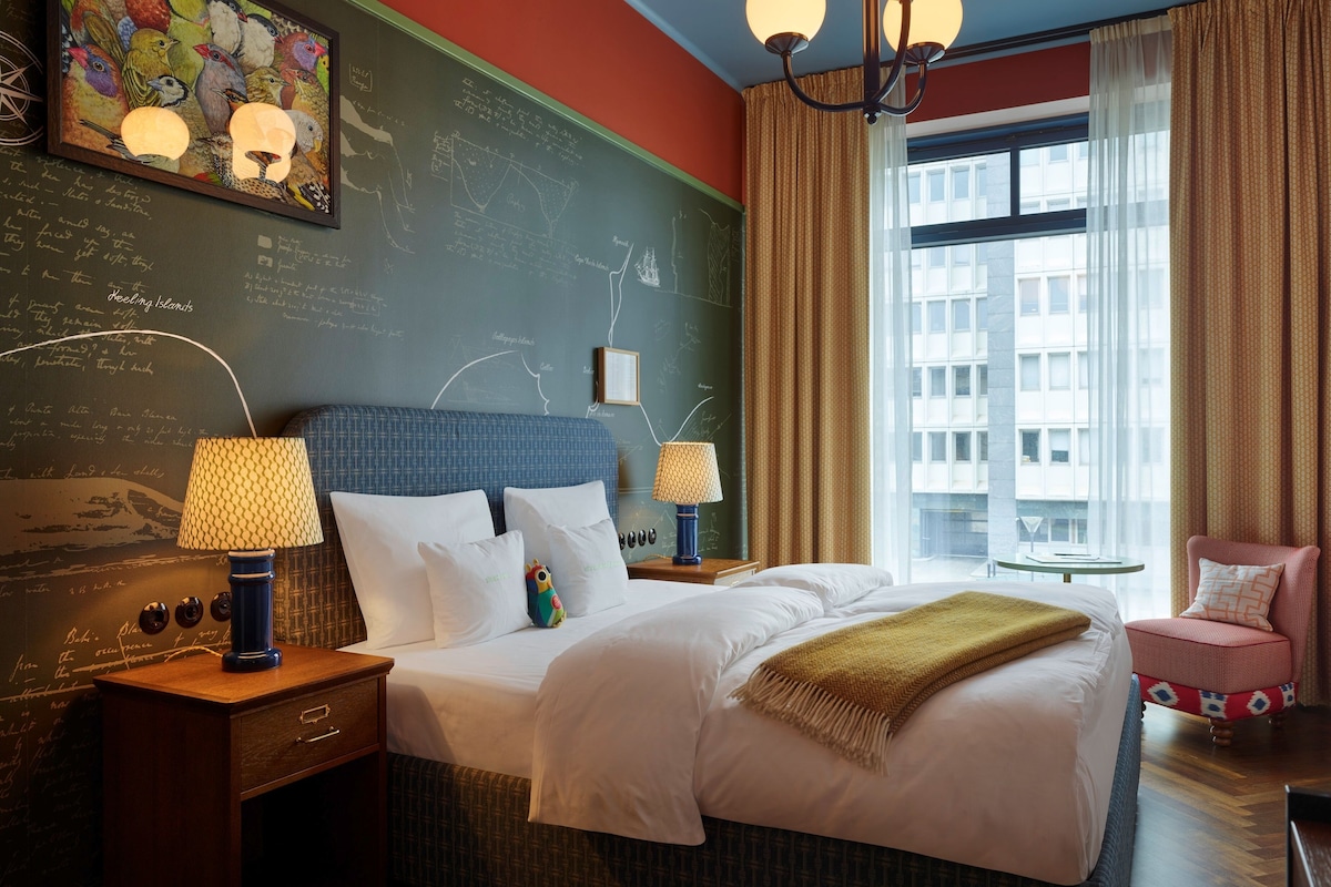 Habitación de hotel con una cama grande, dos mesitas de noche, un ventanal con cortinas, una pared con una pizarra y una pequeña mesa con lámpara.