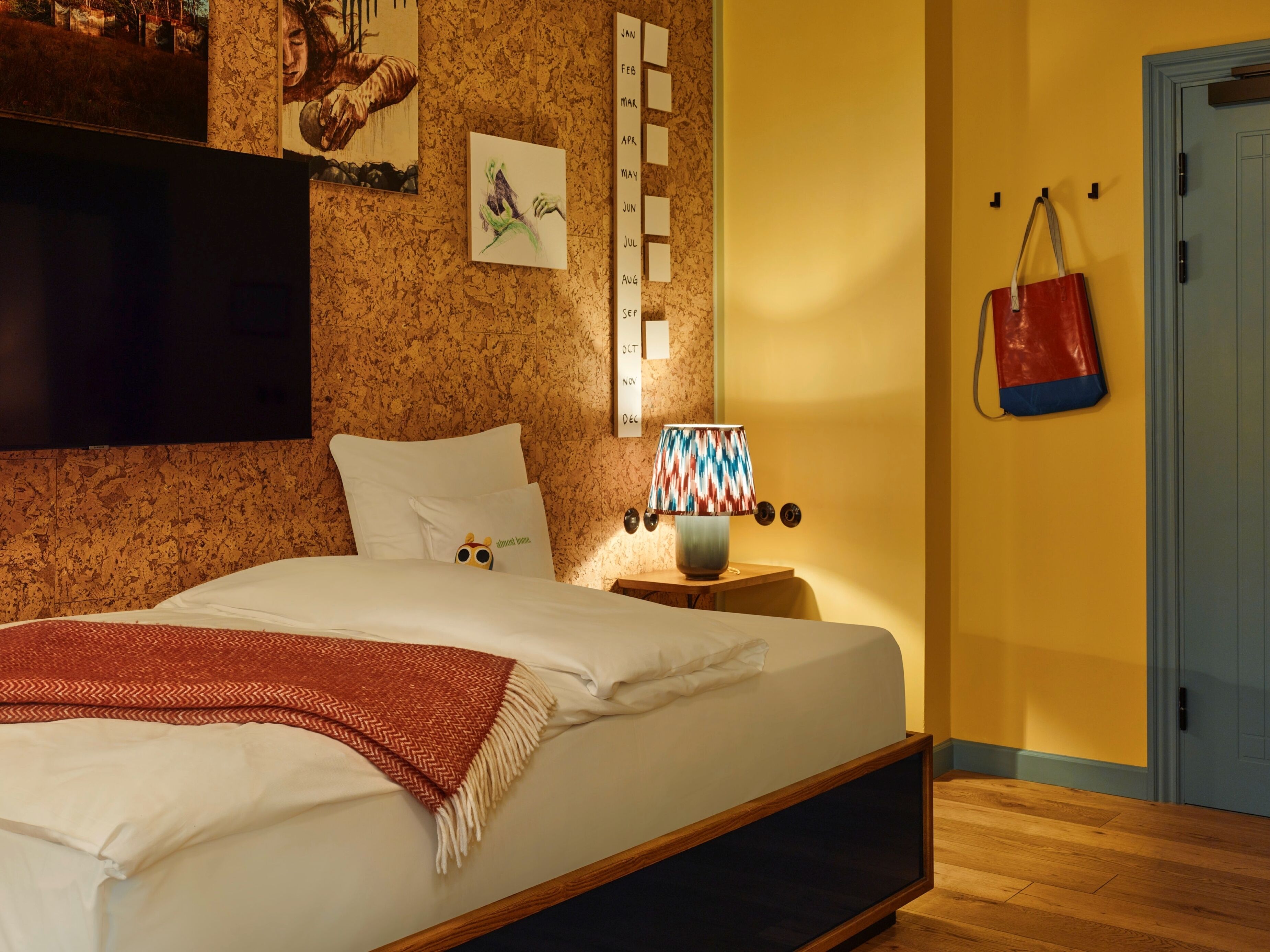 Un dormitorio con cama, televisor de pared, mesita de noche con lámpara, calendario y puerta con un bolso colgado.