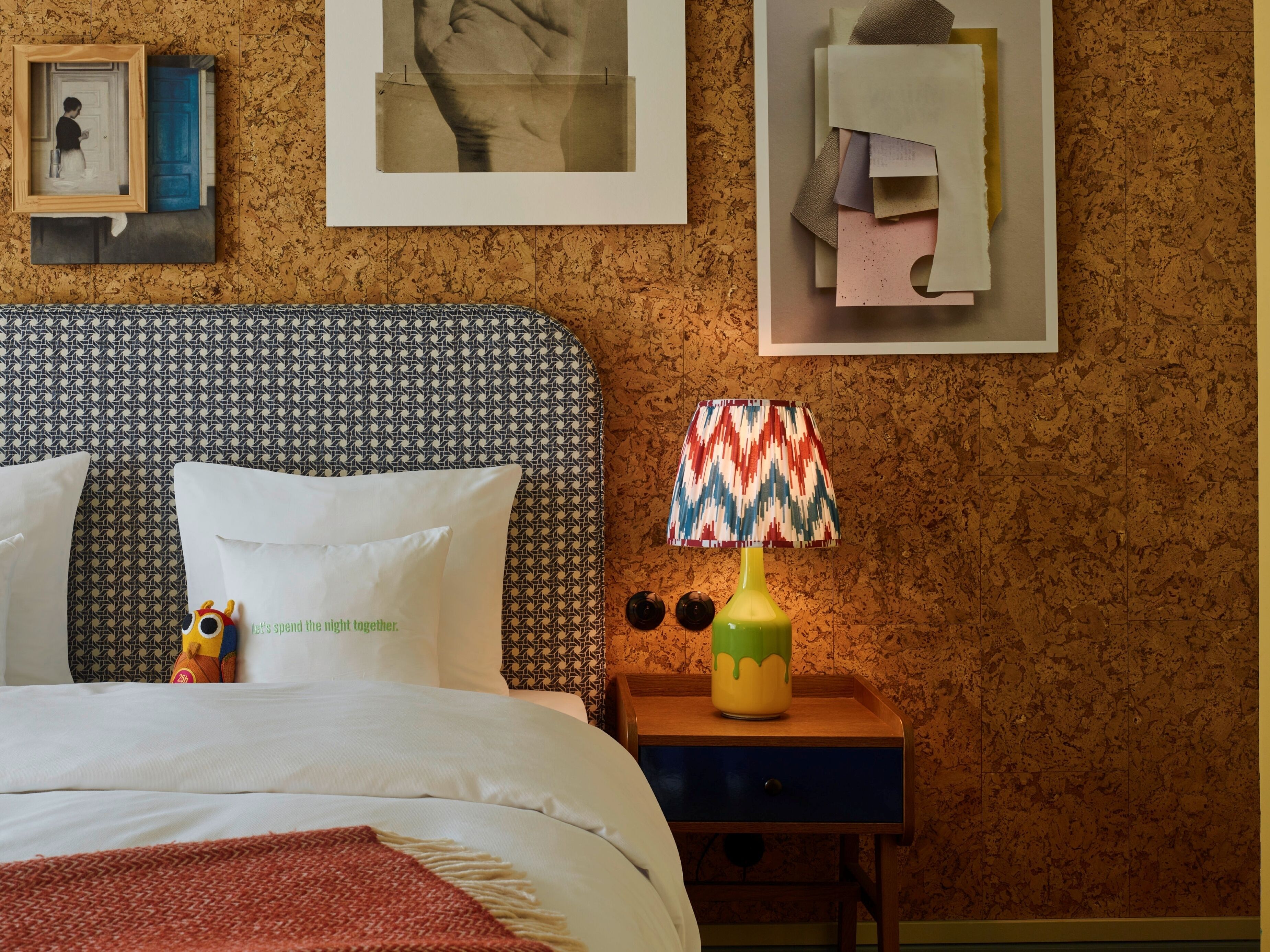 Quarto com cabeceira estampada, cama com roupa de cama branca, luminária colorida sobre uma mesa de cabeceira de madeira e obras de arte nas paredes.