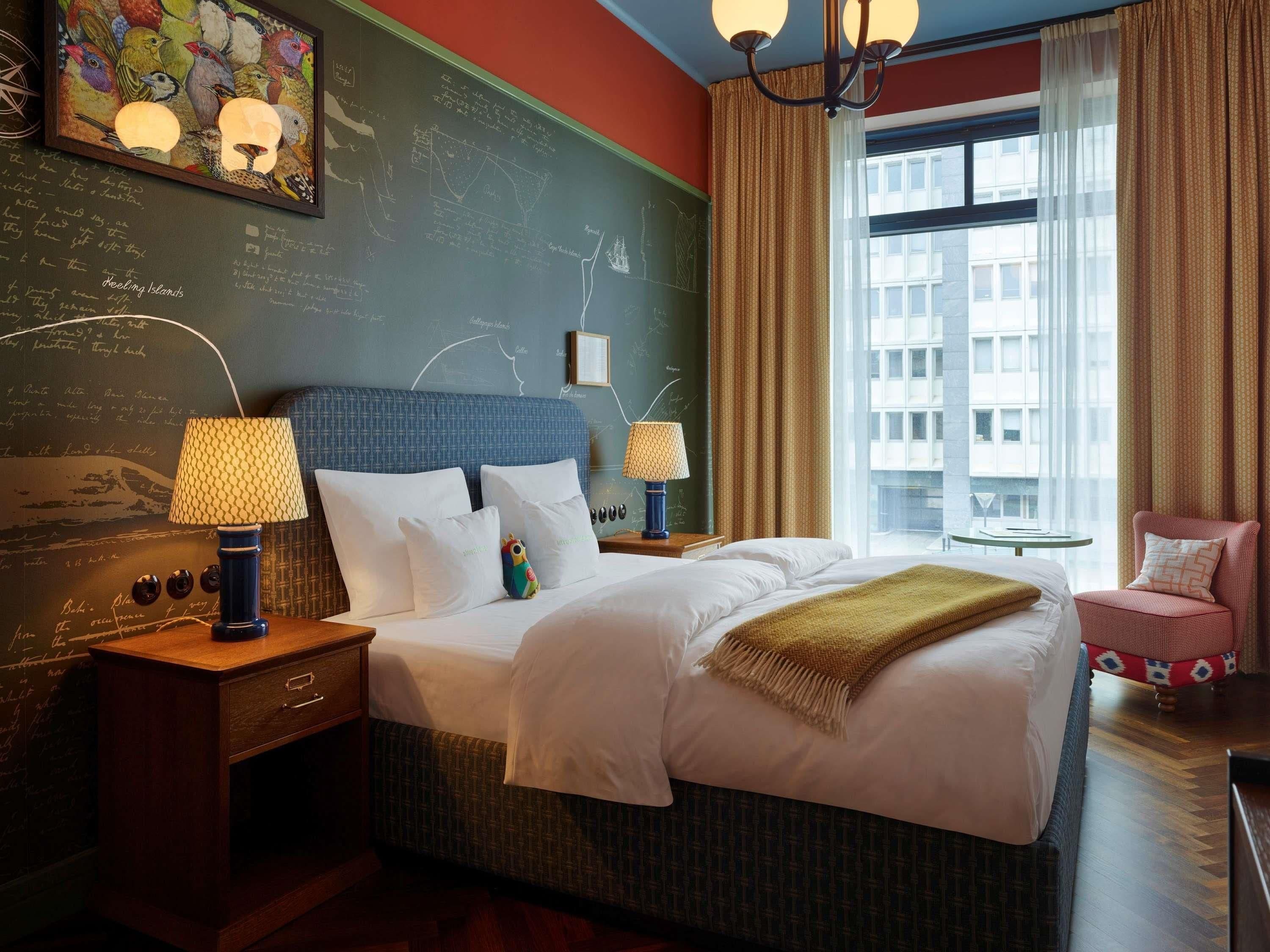 Habitación de hotel con una cama grande, dos mesitas de noche con lámparas, una pequeña mesa con una silla, un ventanal con cortinas y una pared con una pizarra y obras de arte.