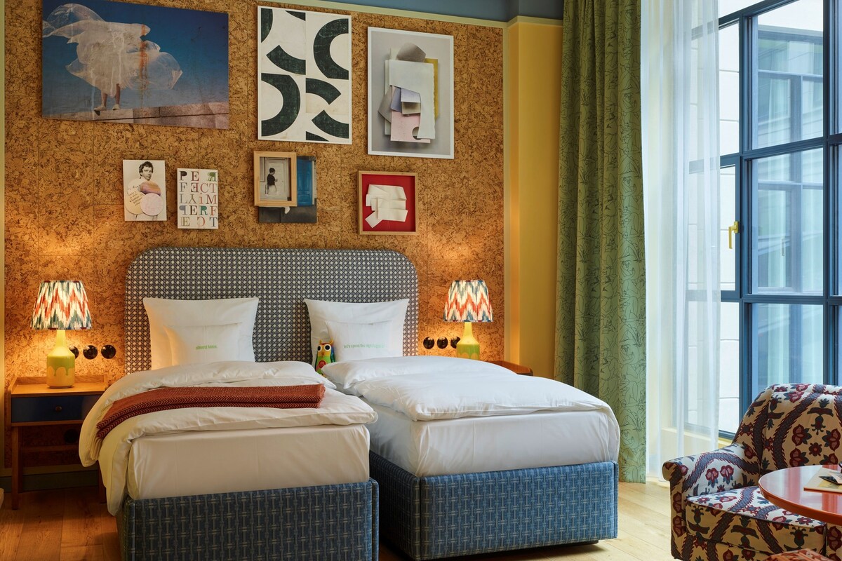 Una cama doble con un cabecero estampado, dos mesitas de noche con lámparas, un sillón con estampado y un ventanal con cortinas.