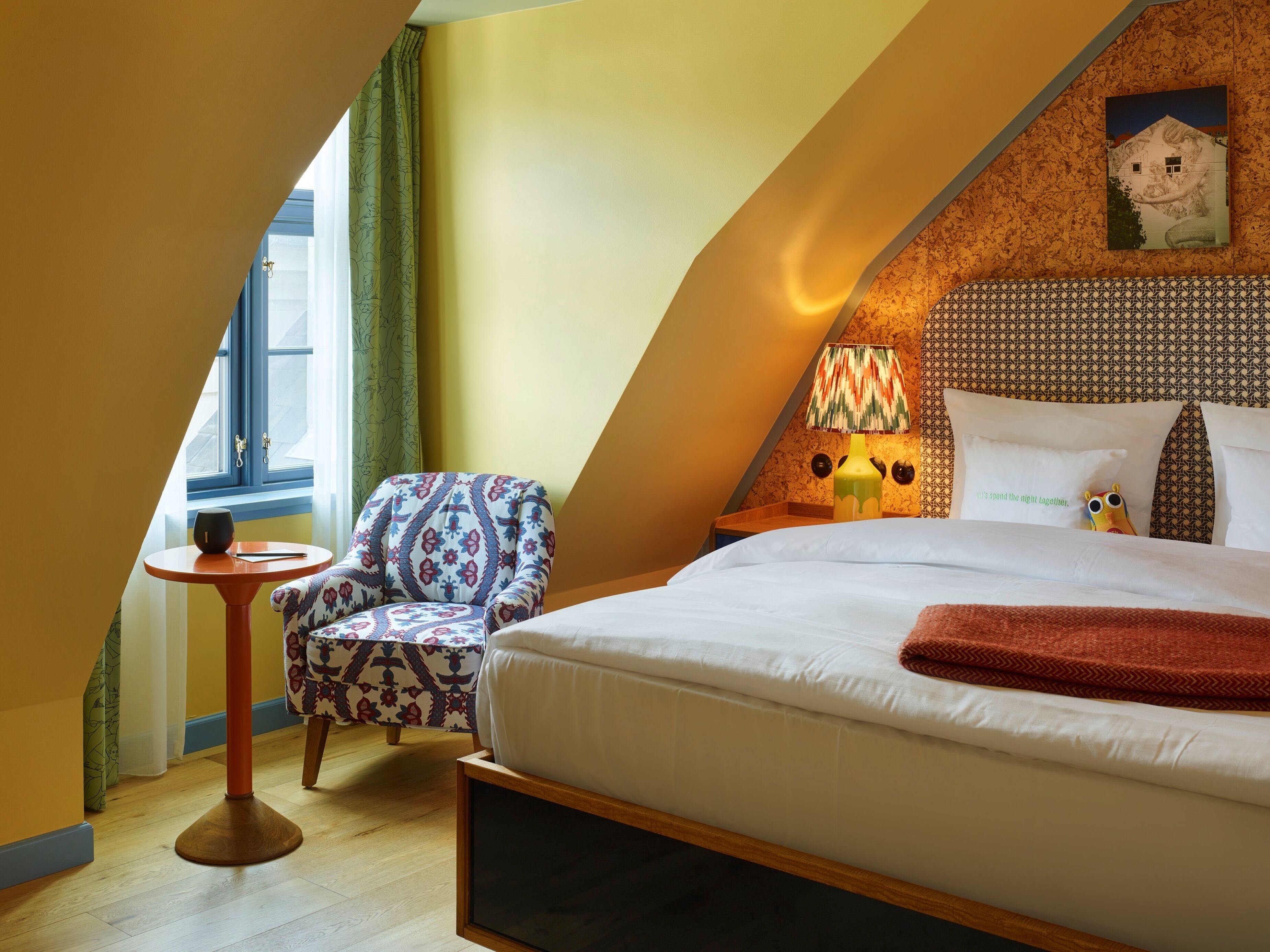 Una camera da letto con un letto, una sedia, un tavolino, una lampada e un quadro appeso al muro.