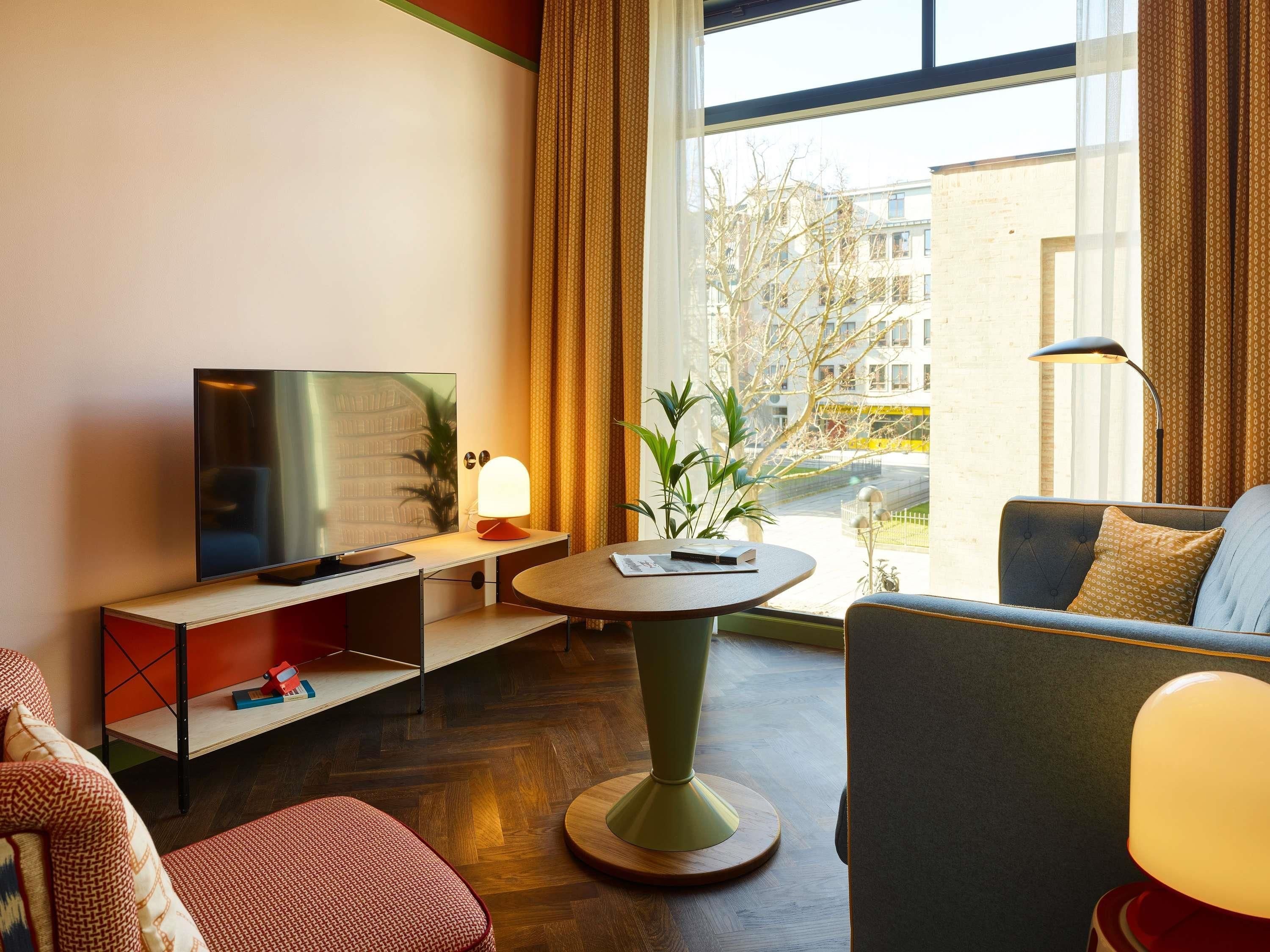 Une chambre d’hôtel moderne équipée d’un téléviseur à écran plat, d’une table ronde avec une lampe, d’un canapé et d’une plante en pot.