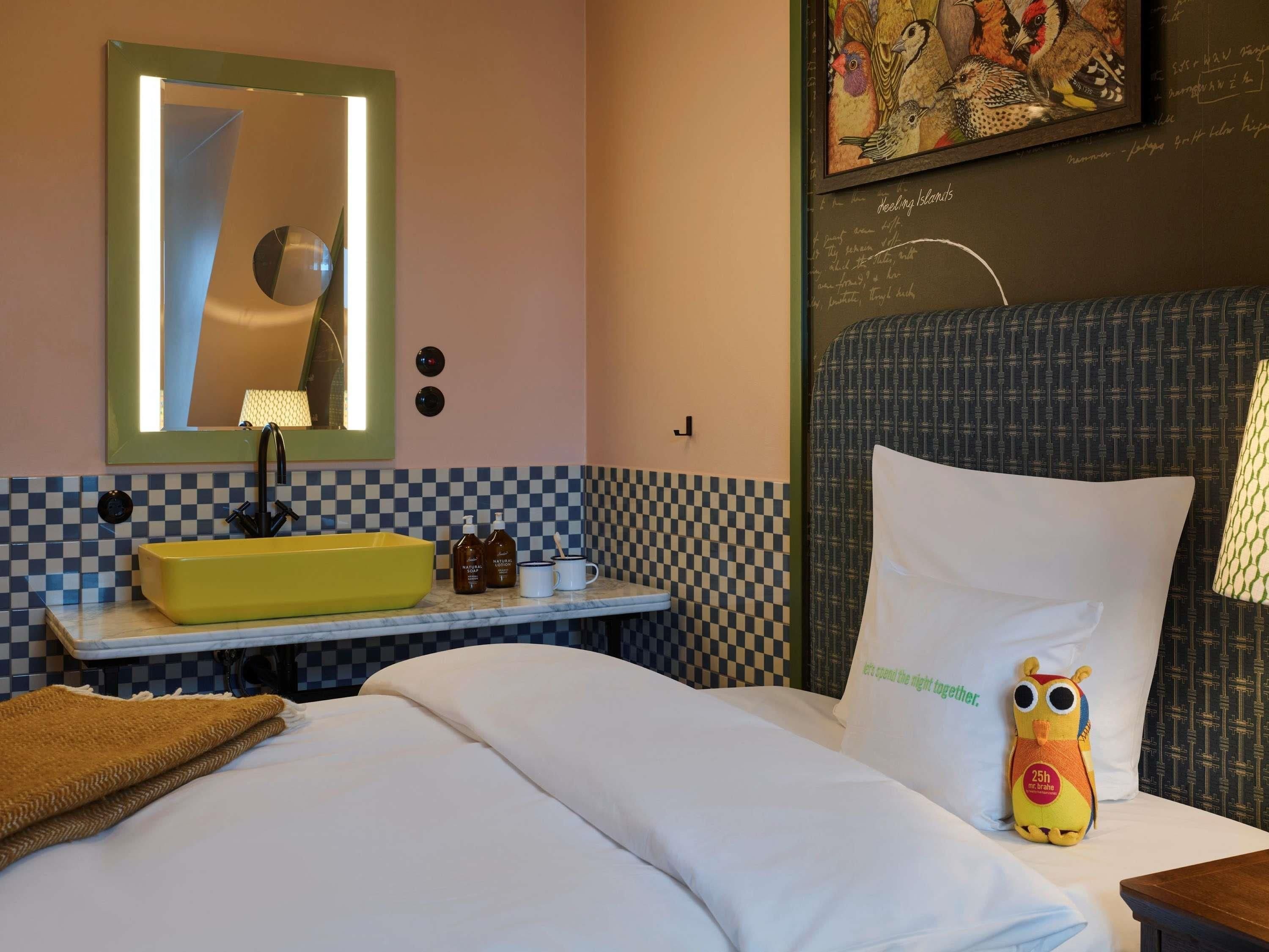 A hotel room with a checkered wall pattern, a yellow sink, a mirror, a bed with white bedding, and a plush toy on the pillow.