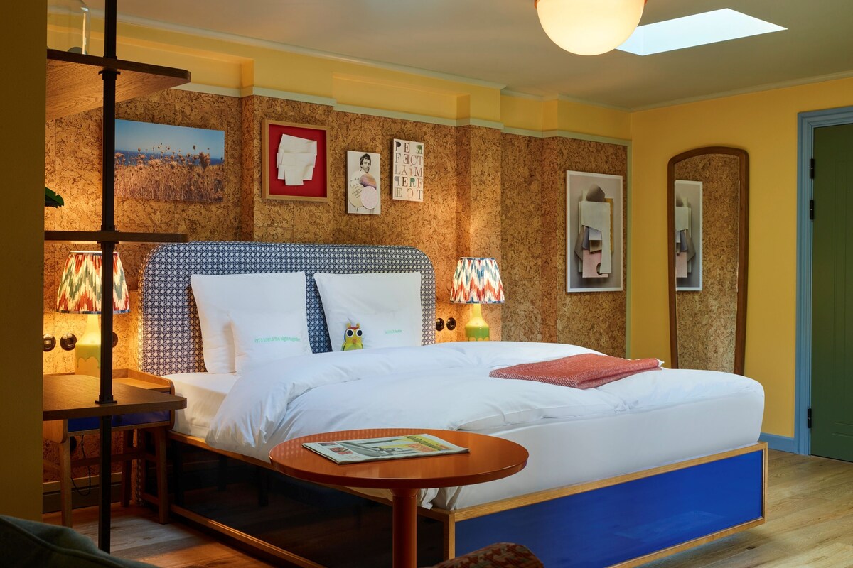 Habitación de hotel con cama, mesita de noche, lámpara y una pequeña mesa redonda con una revista.