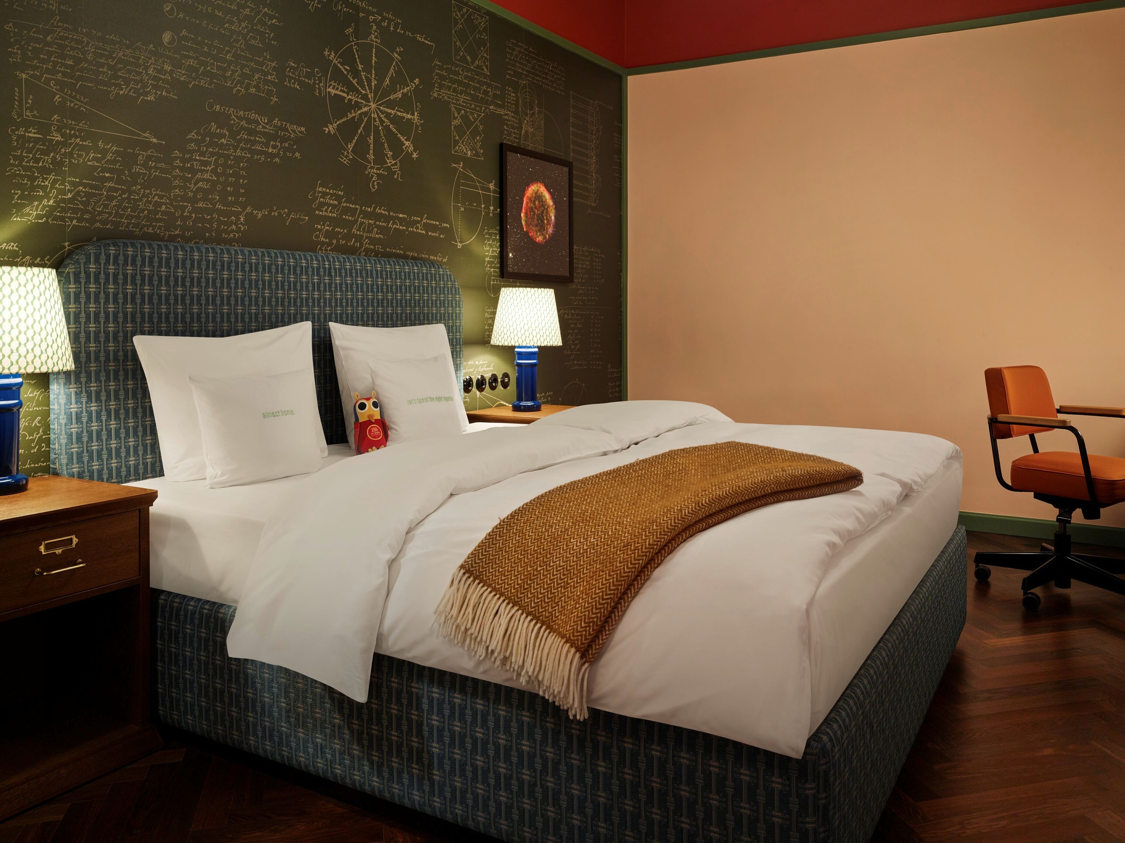 A neatly made bed with a patterned headboard, a nightstand with a lamp, and a wall with scientific diagrams.