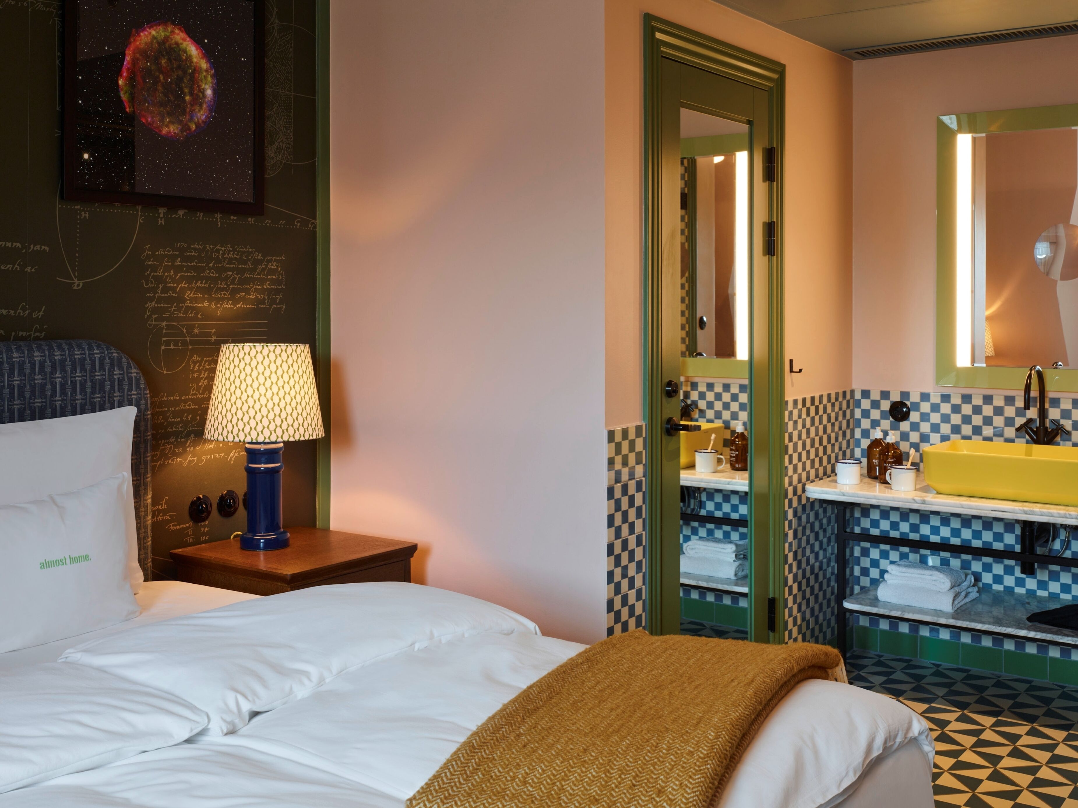Habitación de hotel con cama, mesita de noche con lámpara, baño con lavabo y espejo, y piso a cuadros.
