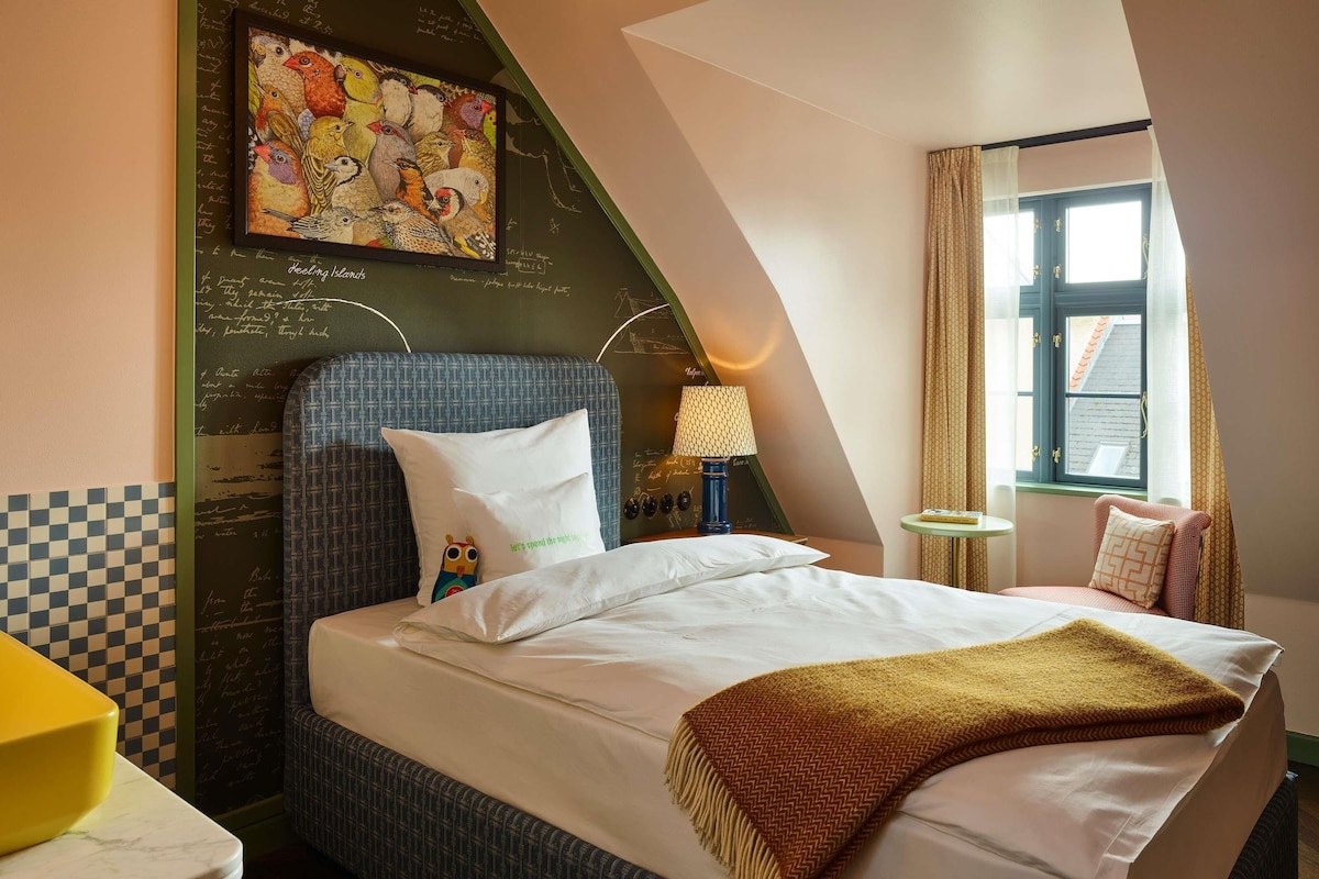 Una habitación de hotel con una cama, un almohadón con diseño de pájaro, una lámpara, una ventana con cortinas y una pared con una pizarra.