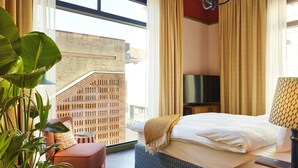 Una habitación de hotel con una cama, televisor, planta y vistas a un edificio de ladrillo.
