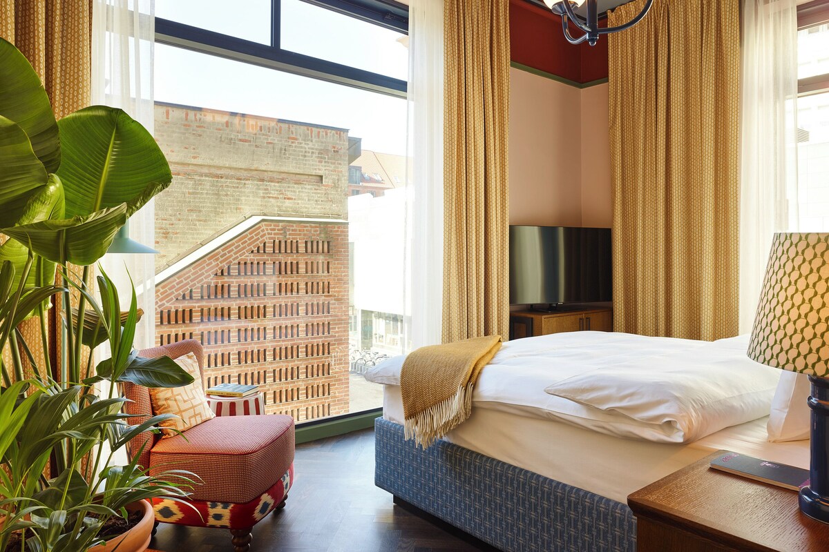 Una habitación de hotel con una cama, televisor, planta y vistas a un edificio de ladrillo.