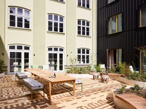 A courtyard with a wooden table and benches, surrounded by buildings with large windows and a garden area.