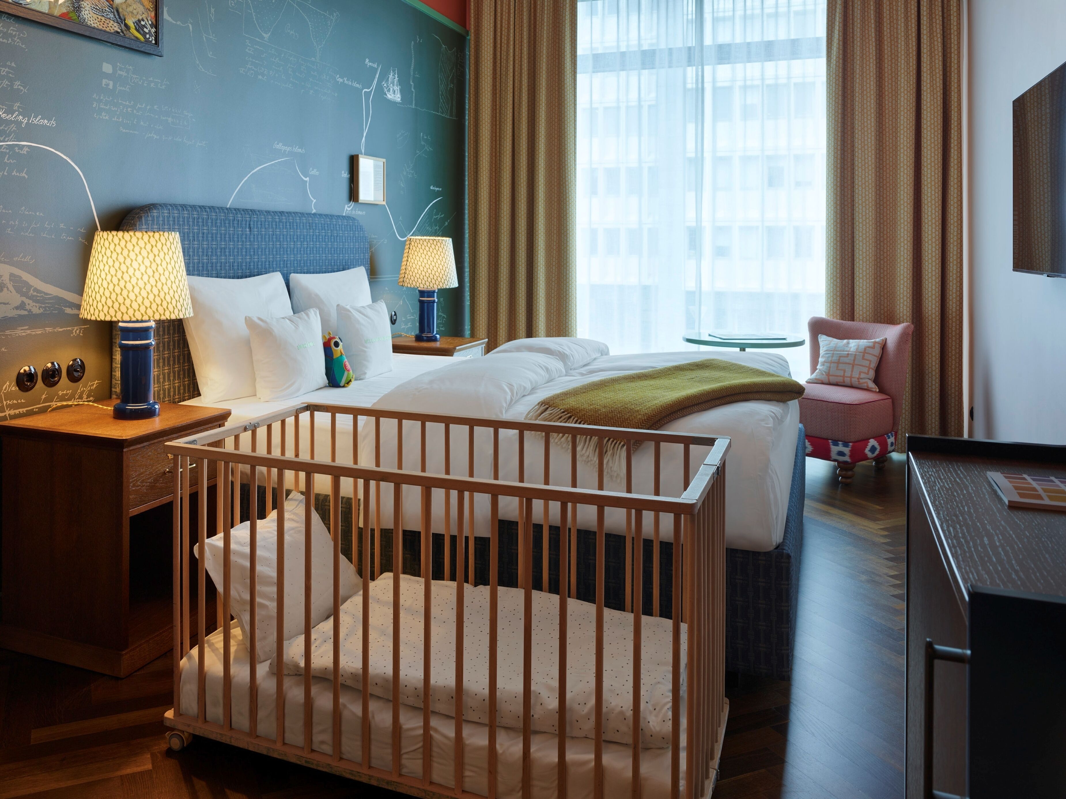 Quarto de hotel com berço, cama, escrivaninha, cadeira, abajur e parede com lousa.
