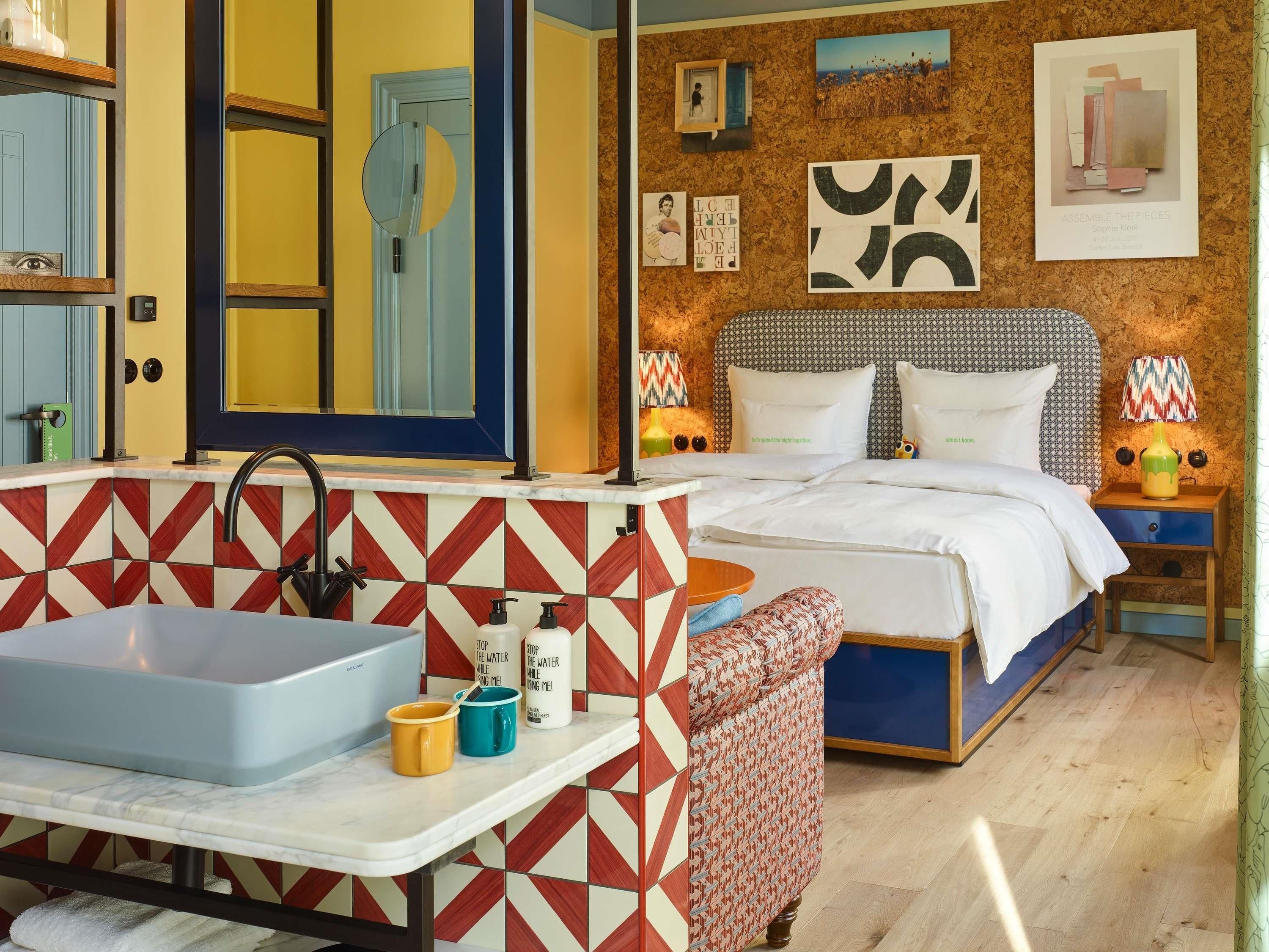 Una habitación de hotel con baño, una cama, una mesita de noche y una pared decorada con obras de arte.