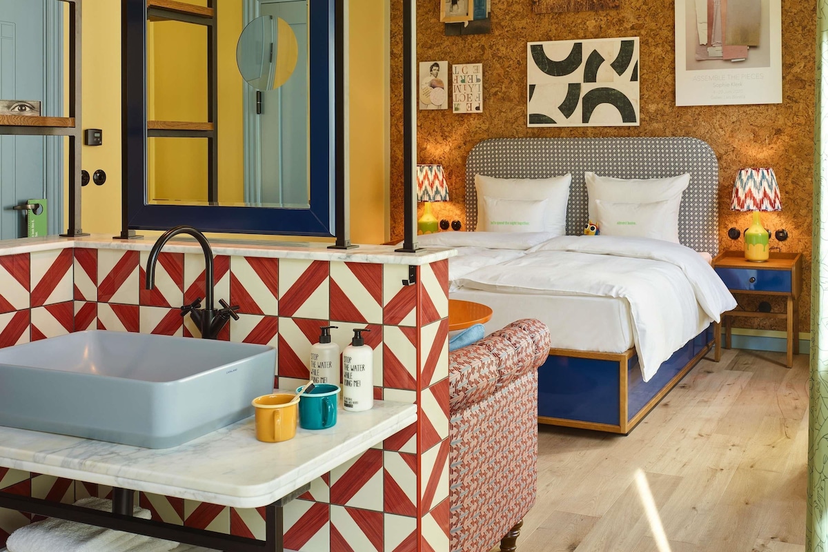 Una habitación de hotel con baño, una cama, una mesita de noche y una pared decorada con obras de arte.
