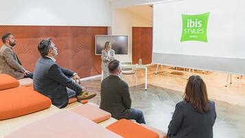 Un salón de conferencias con una pantalla de presentación, una pizarra blanca y el logotipo de ibis Styles Hotels.