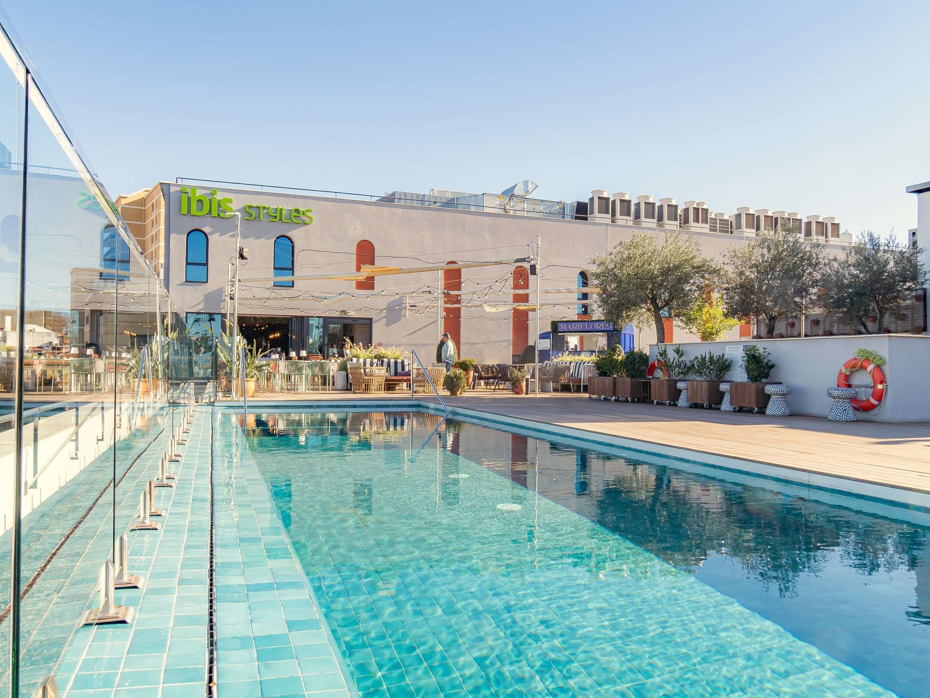 Une piscine avec un bord carrelé, un restaurant appelé « ibis Styles » en arrière-plan, et un espace extérieur aménagé avec des tables et des chaises.