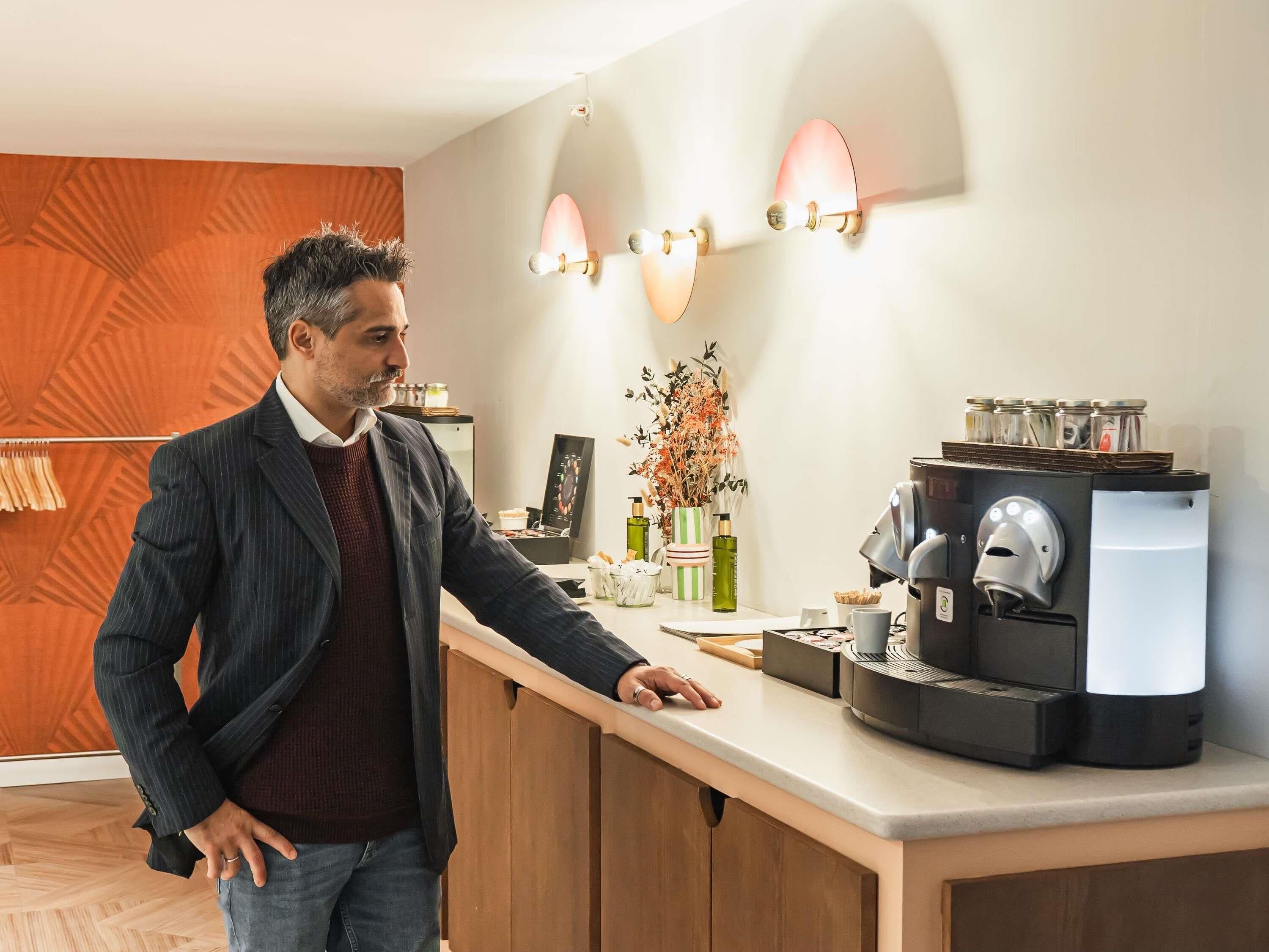 Un hombre con traje parado en un mostrador con una máquina de café, un florero con flores y apliques de pared.