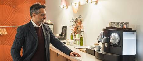 Un hombre con traje parado en un mostrador con una máquina de café, un florero con flores y apliques de pared.