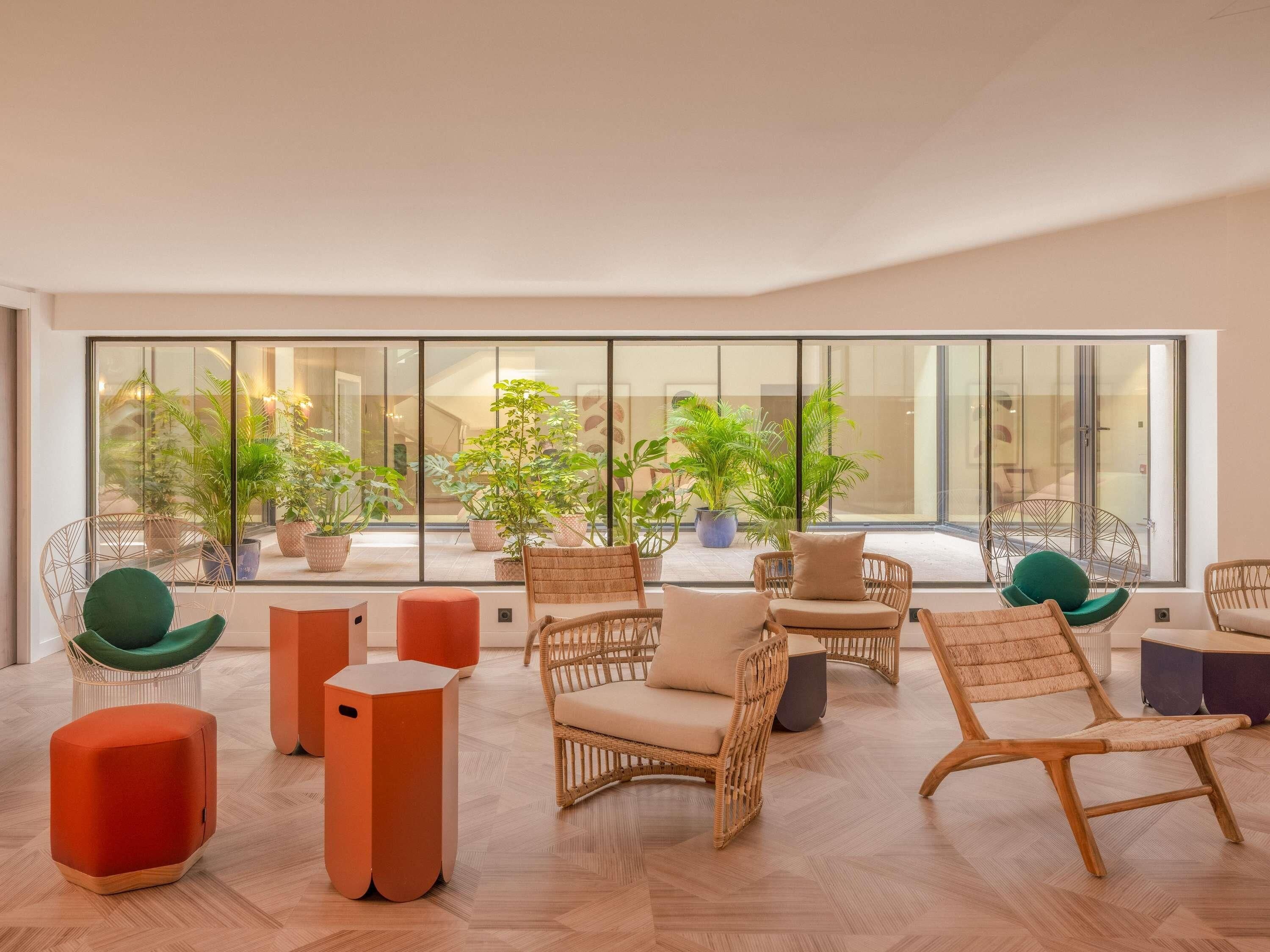 Un espace intérieur vaste et lumineux, aménagé avec des meubles en osier, des plantes en pot et une grande fenêtre offrant une vue sur l’extérieur.