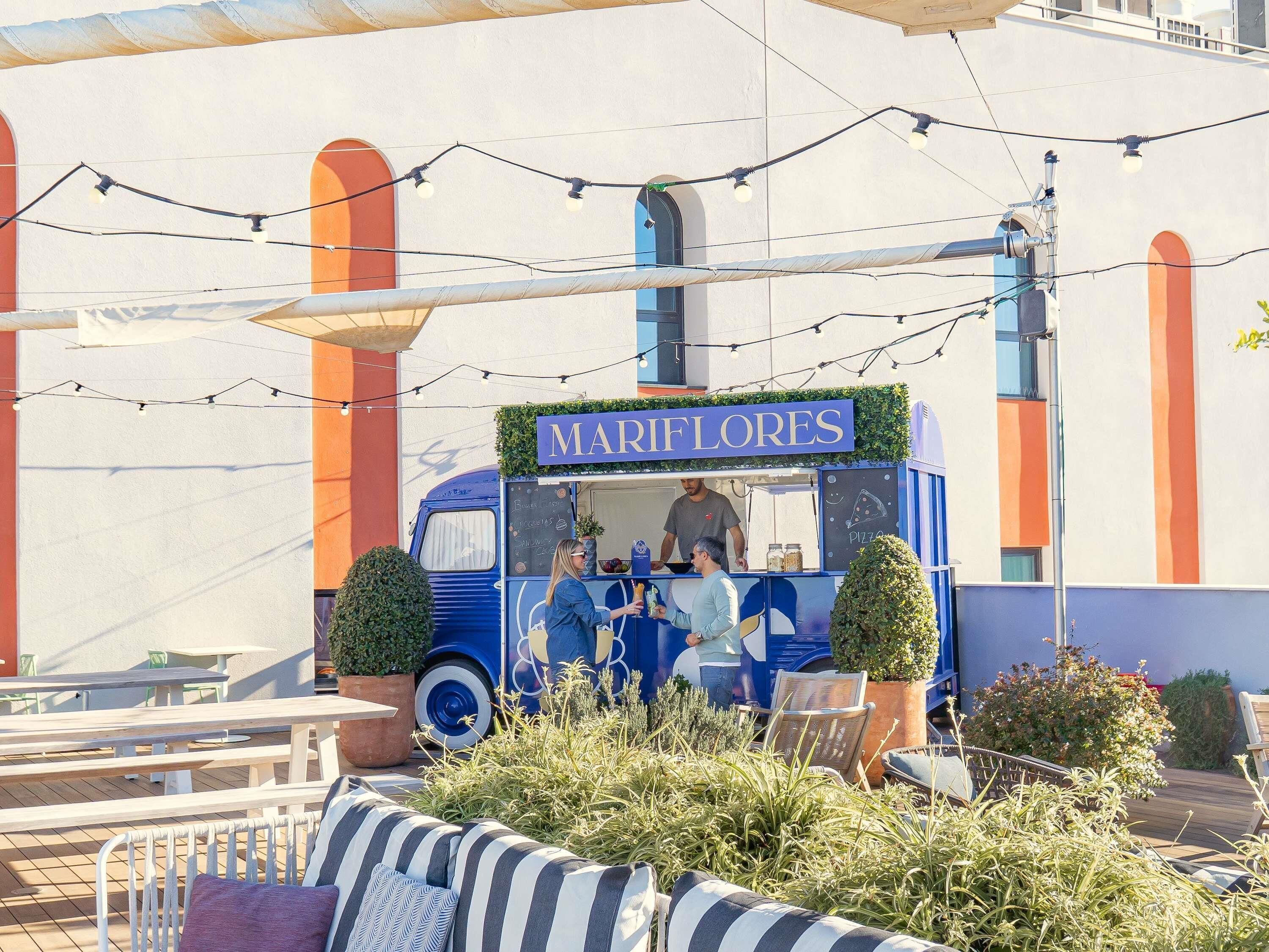 A food truck named 'MARIFLORES' with a blue and white color scheme, serving food, and located in an outdoor seating area with tables and chairs.