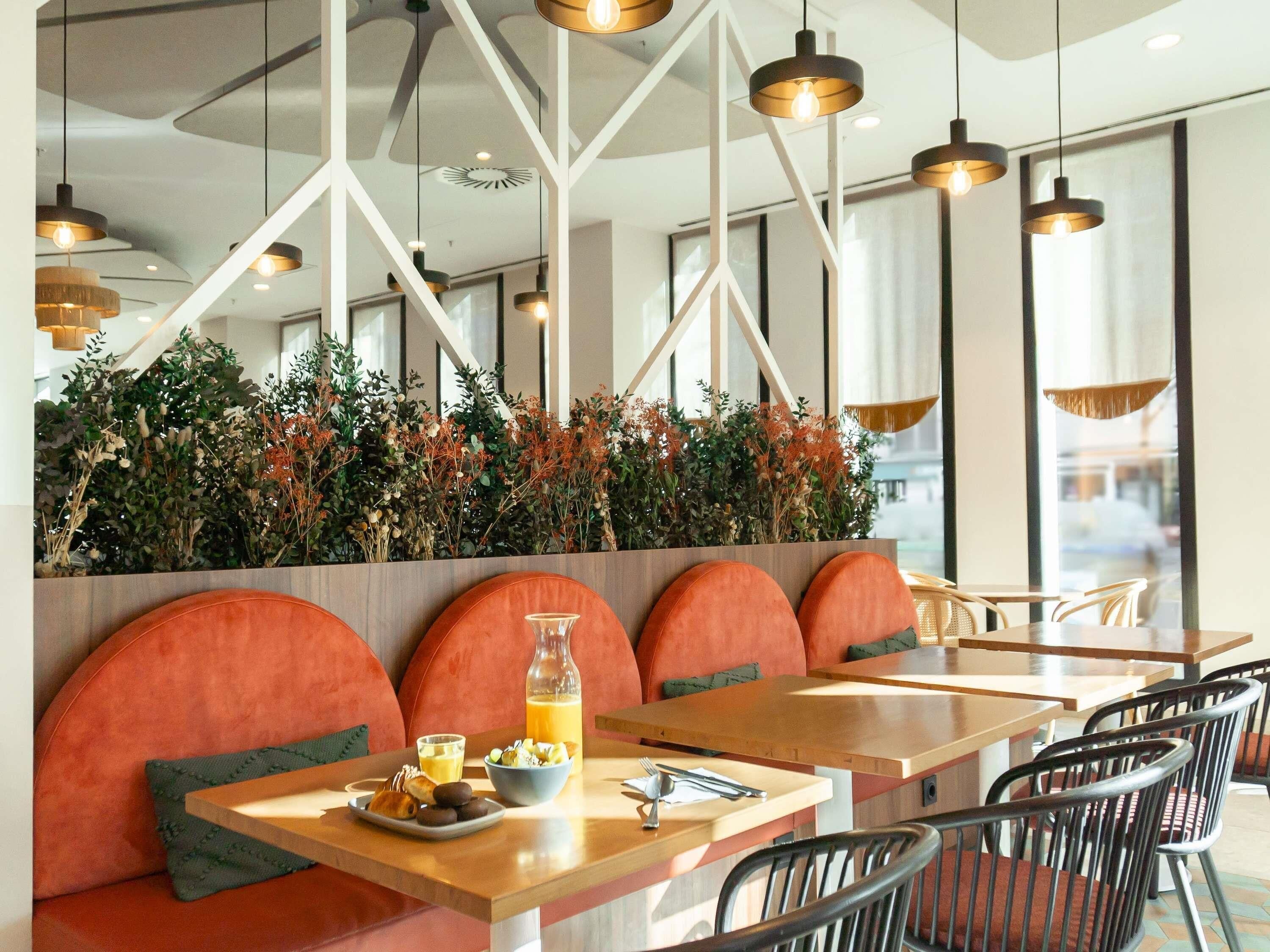 A modern restaurant interior with wooden tables, orange cushioned seating, and hanging pendant lights.