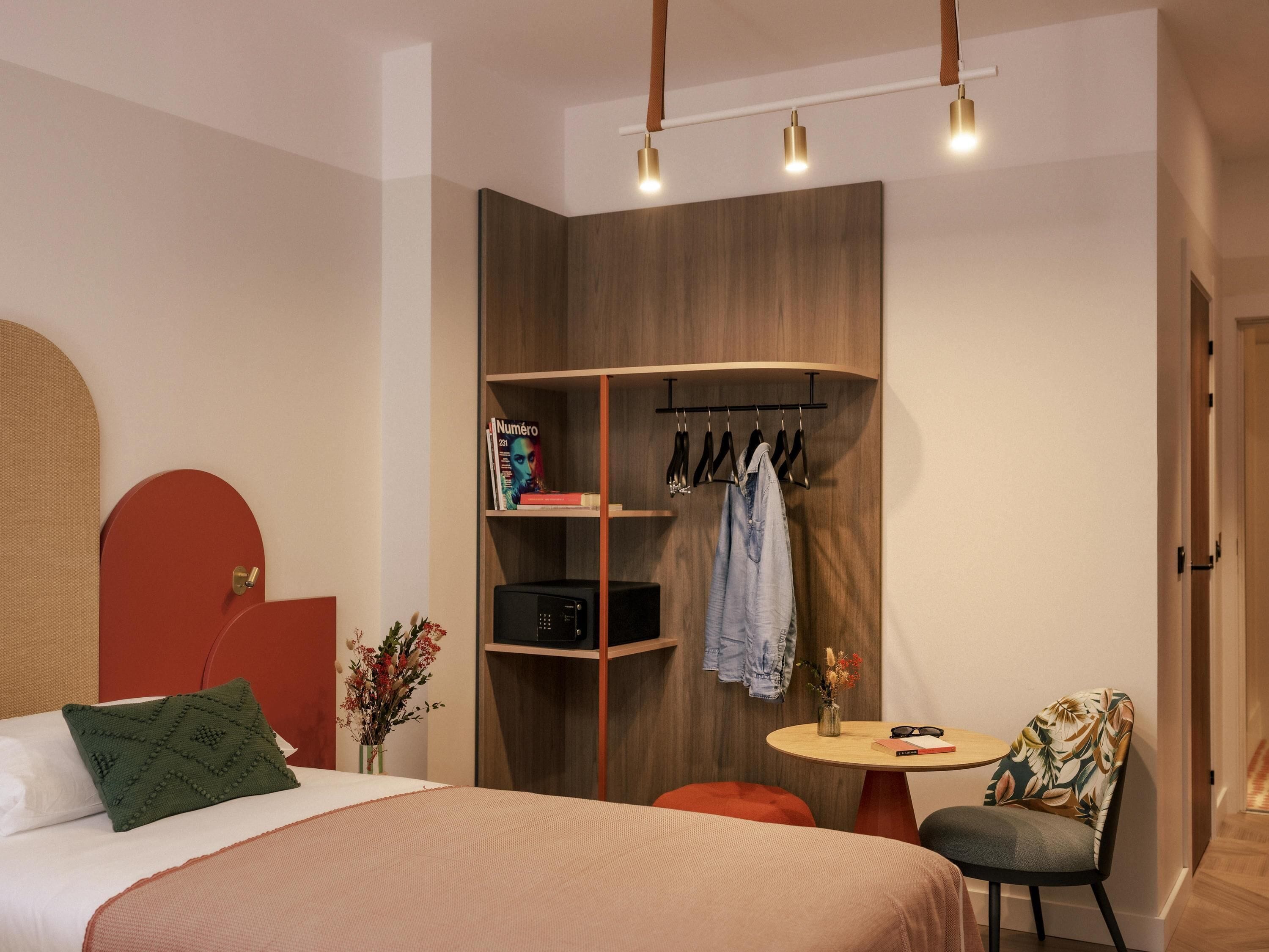 Un dormitorio moderno con cama, una mesita redonda pequeña, una silla, un armario y una percha con ropa.