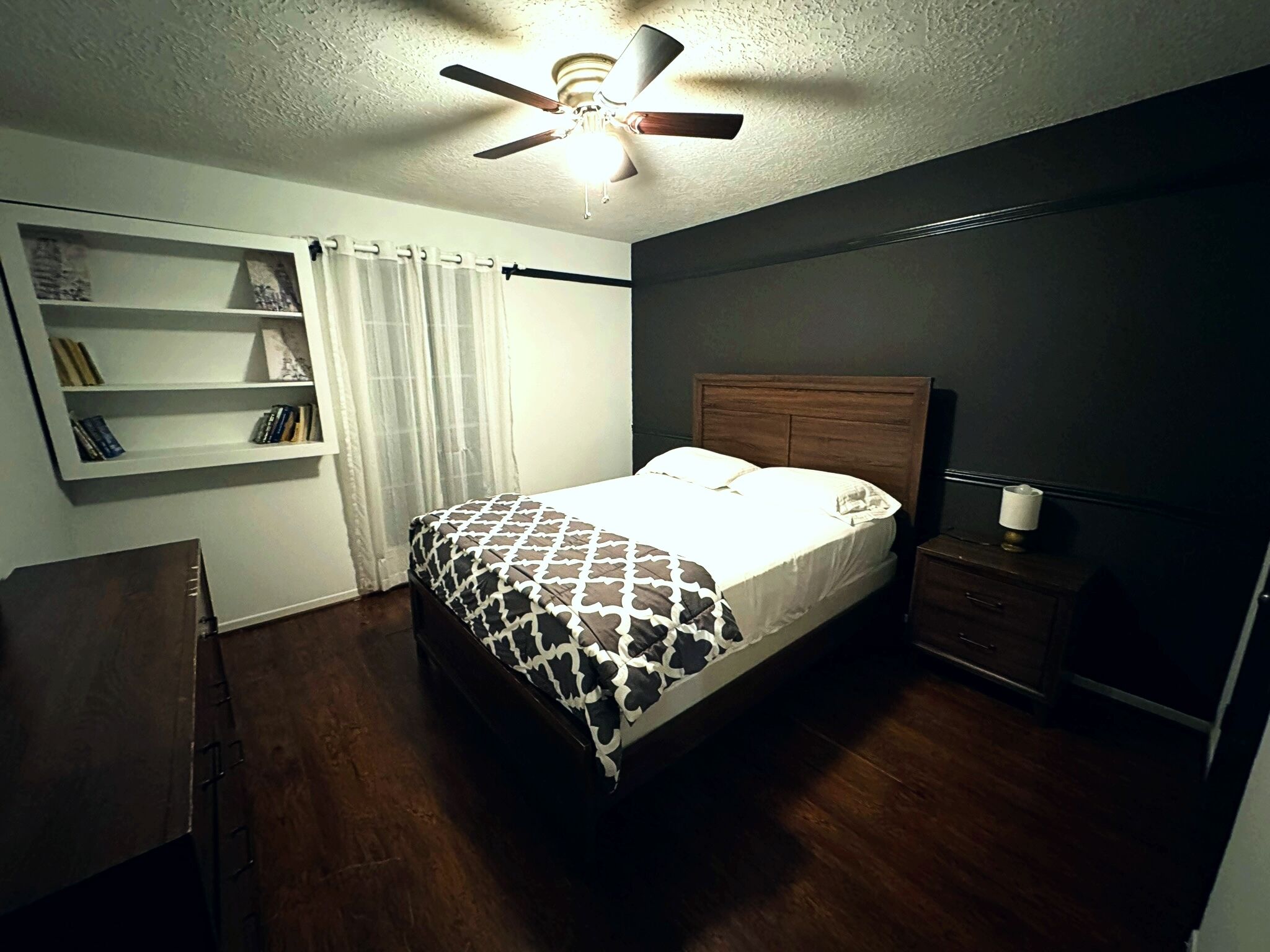 A bedroom with a bed, a ceiling fan, a bedside table, a nightstand, a bookshelf, and a window with curtains.