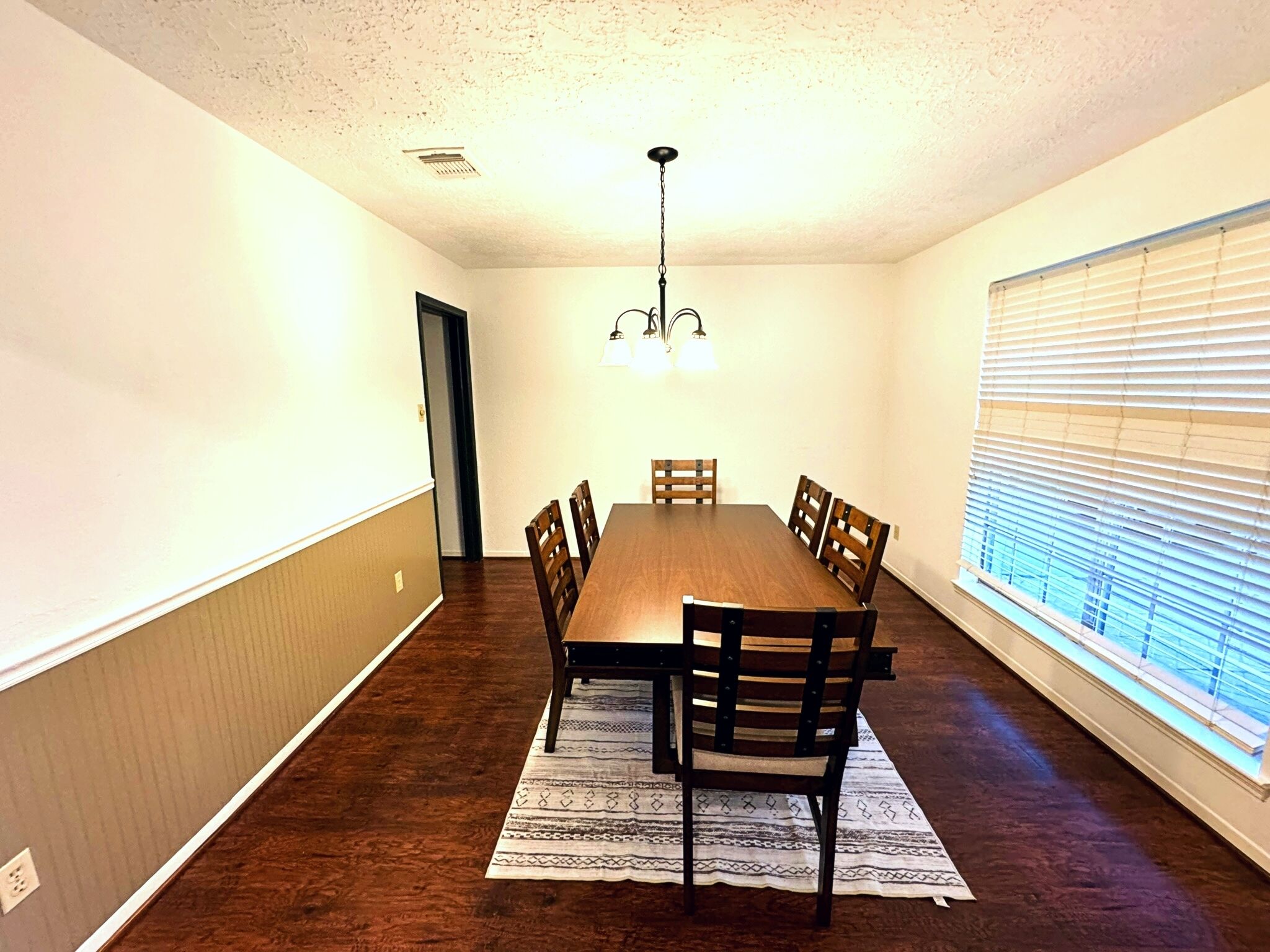 A dining room with a wooden table and chairs, a patterned rug, and a ceiling light fixture.