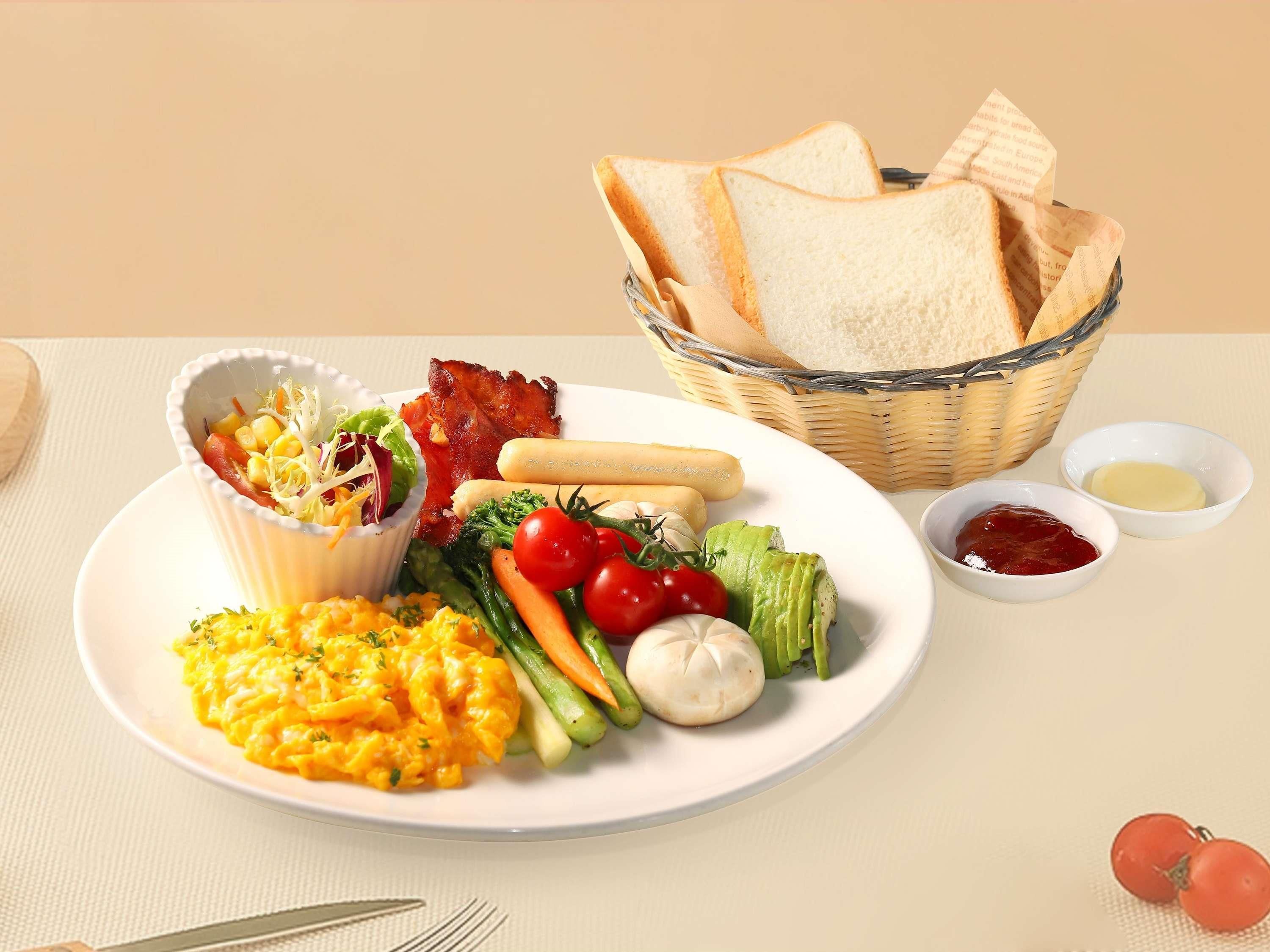 A plate with scrambled eggs, bacon, vegetables, and bread rolls.