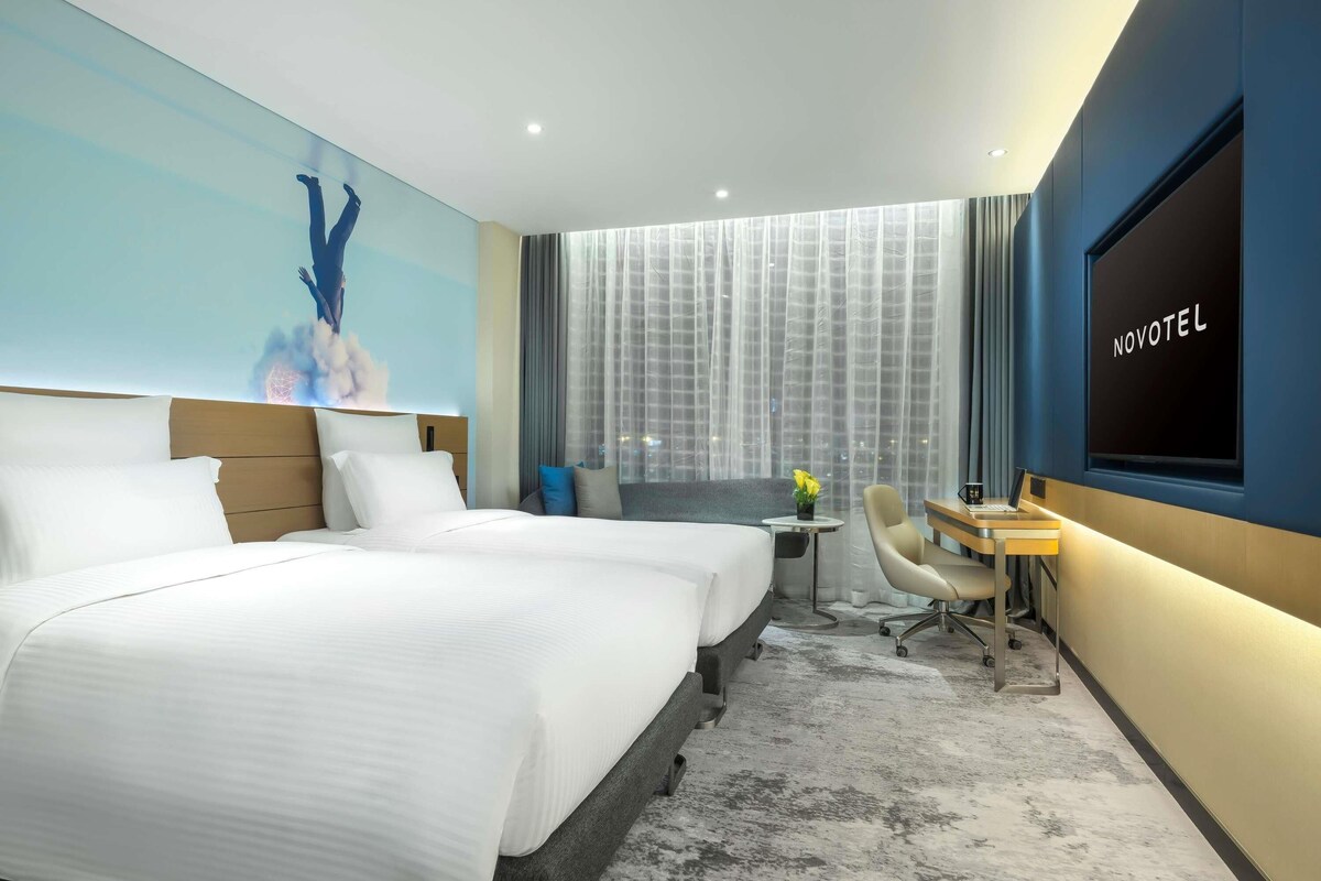 A hotel room with two beds, a desk, a chair, a TV, and a large window with curtains.