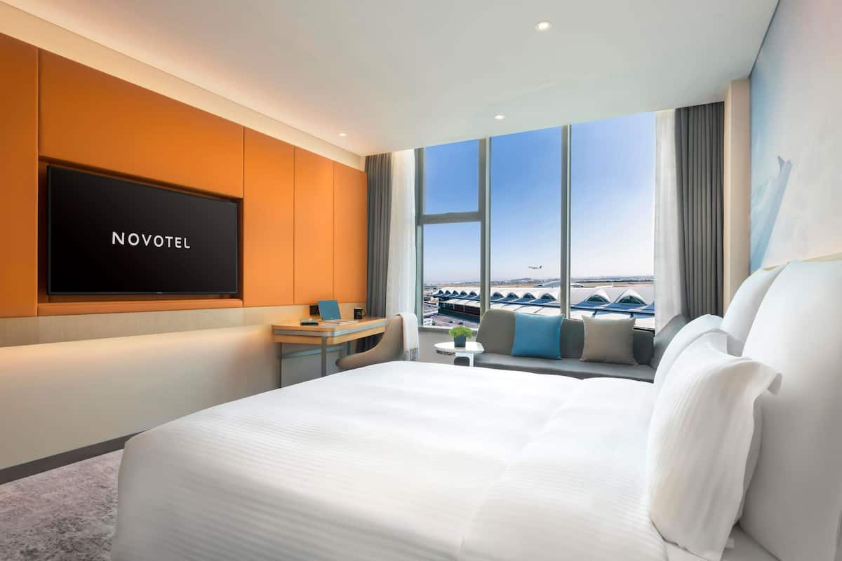 A hotel room with a bed, a desk with a laptop, a TV displaying 'NOVOTEL,' and a view of an airport.