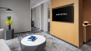 Een hotel receptie met een receptiebalie, een flatscreen televisie waarop 'NOVOTEL' staat, een ronde salontafel met boeken, een vaas met gele bloemen en een moderne lamp.