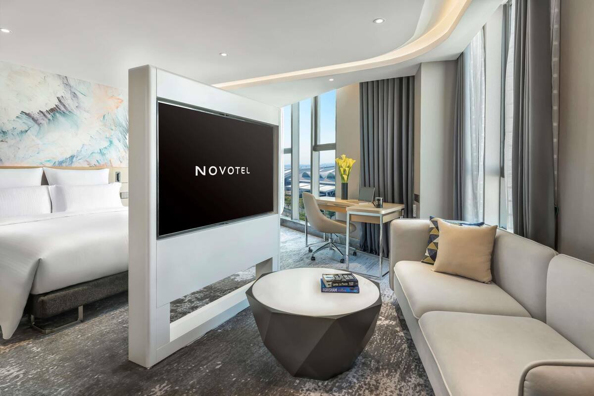 A modern hotel room with a large flat-screen TV, a sofa, a coffee table, and a dining area with a view.