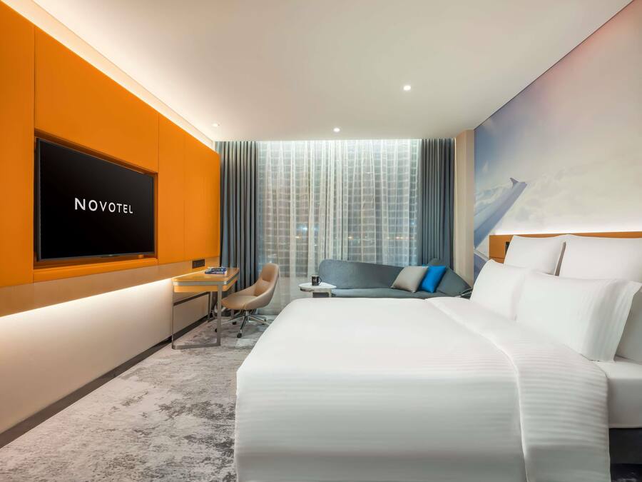 A hotel room with a bed, a desk, a chair, a TV, and a large window with curtains.