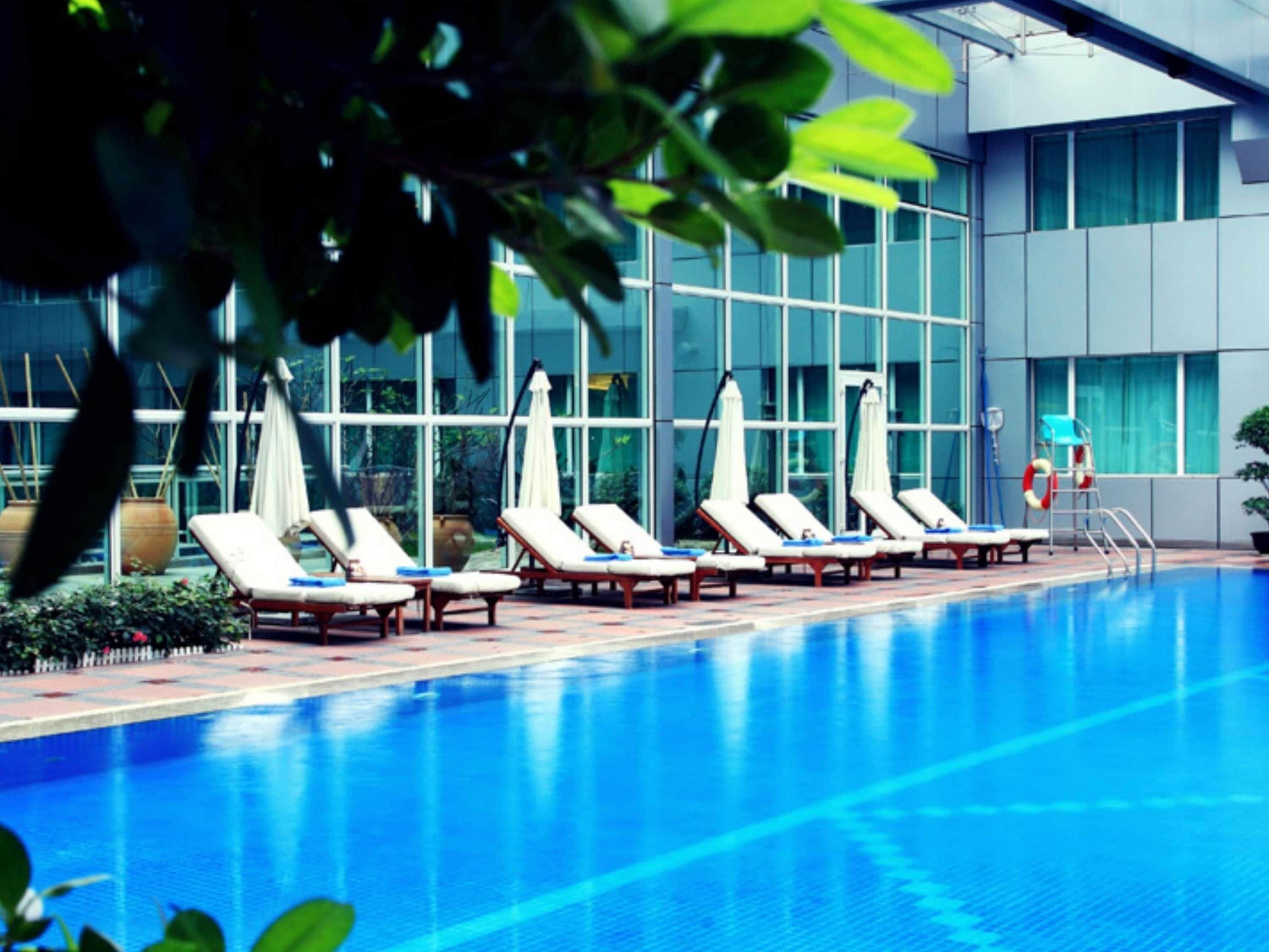 Ein Swimmingpool mit Liegestühlen und Sonnenschirmen, umgeben von einem Gebäude mit großen Fenstern.