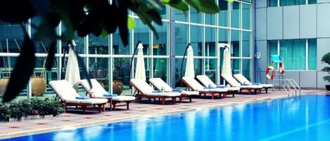 Una piscina con sillas de descanso y sombrillas, rodeada por un edificio con grandes ventanales.