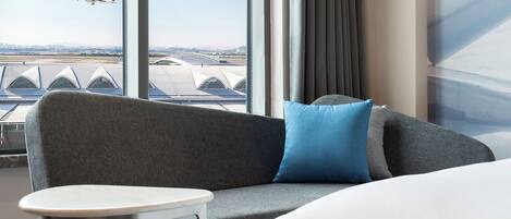 A modern hotel room with a grey sofa, a small round table, and a view of an airport with an airplane in the distance.