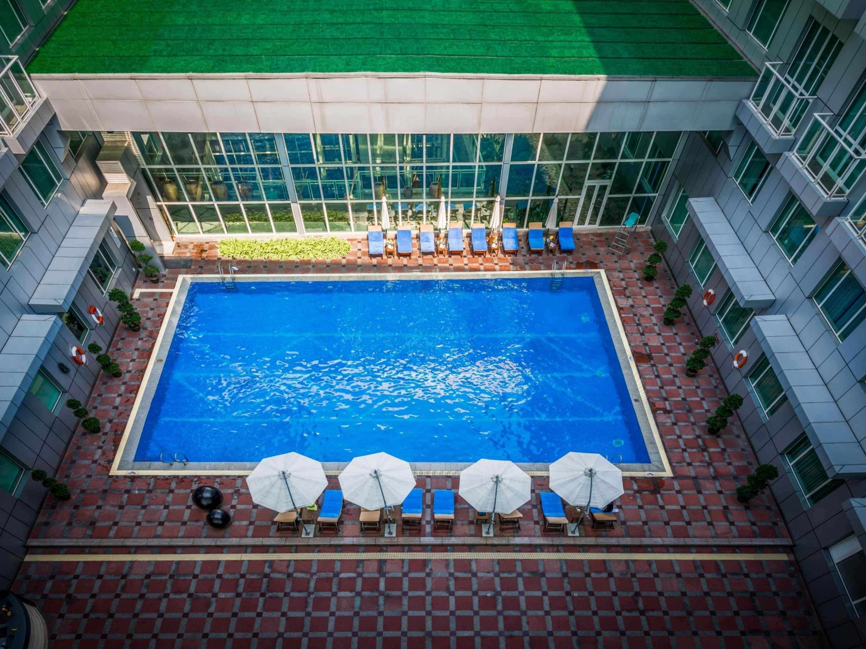 Piscina en la azotea con tumbonas y sombrillas, rodeada por un edificio moderno con grandes ventanales.